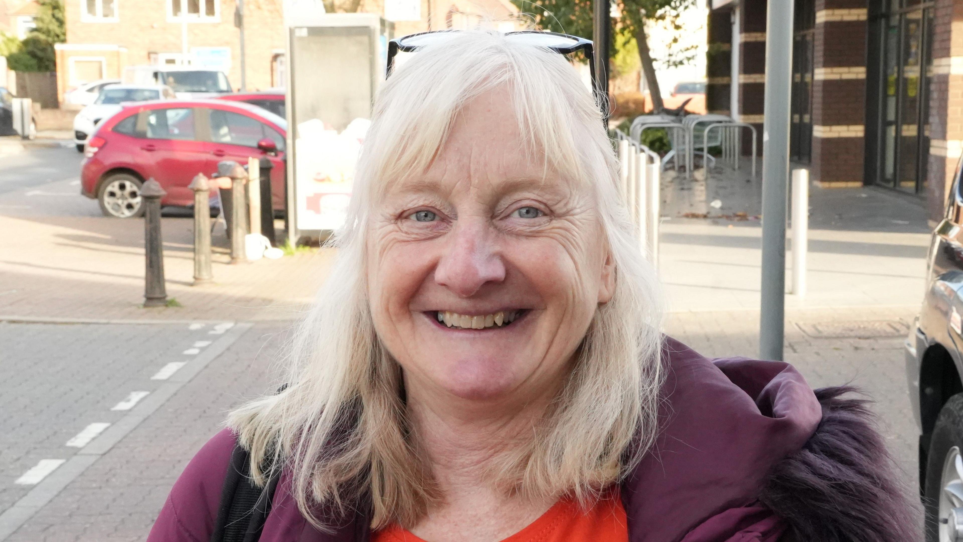 Joanne Ovenall stands outside a supermarket and smiles at the camera. She has long grey hair with glasses resting on top of her head. She wears a dark pink coat with a red top underneath. Parked cars can be seen behind her.