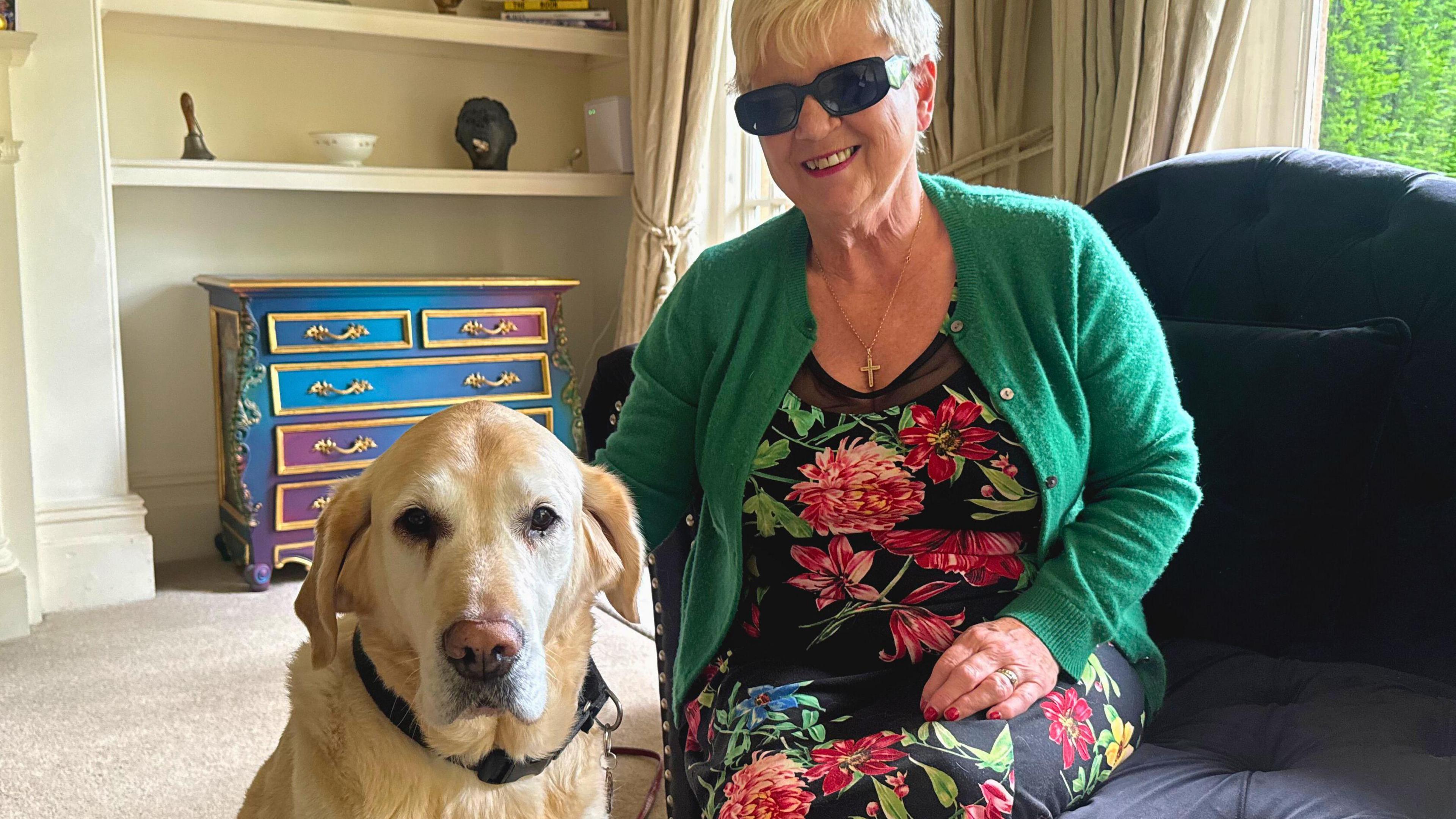 A woman, who wears sunglasses, sits on a dark-coloured sofa and smiles. Beside her is a light-coloured Labrador, wearing a guide dog harness.