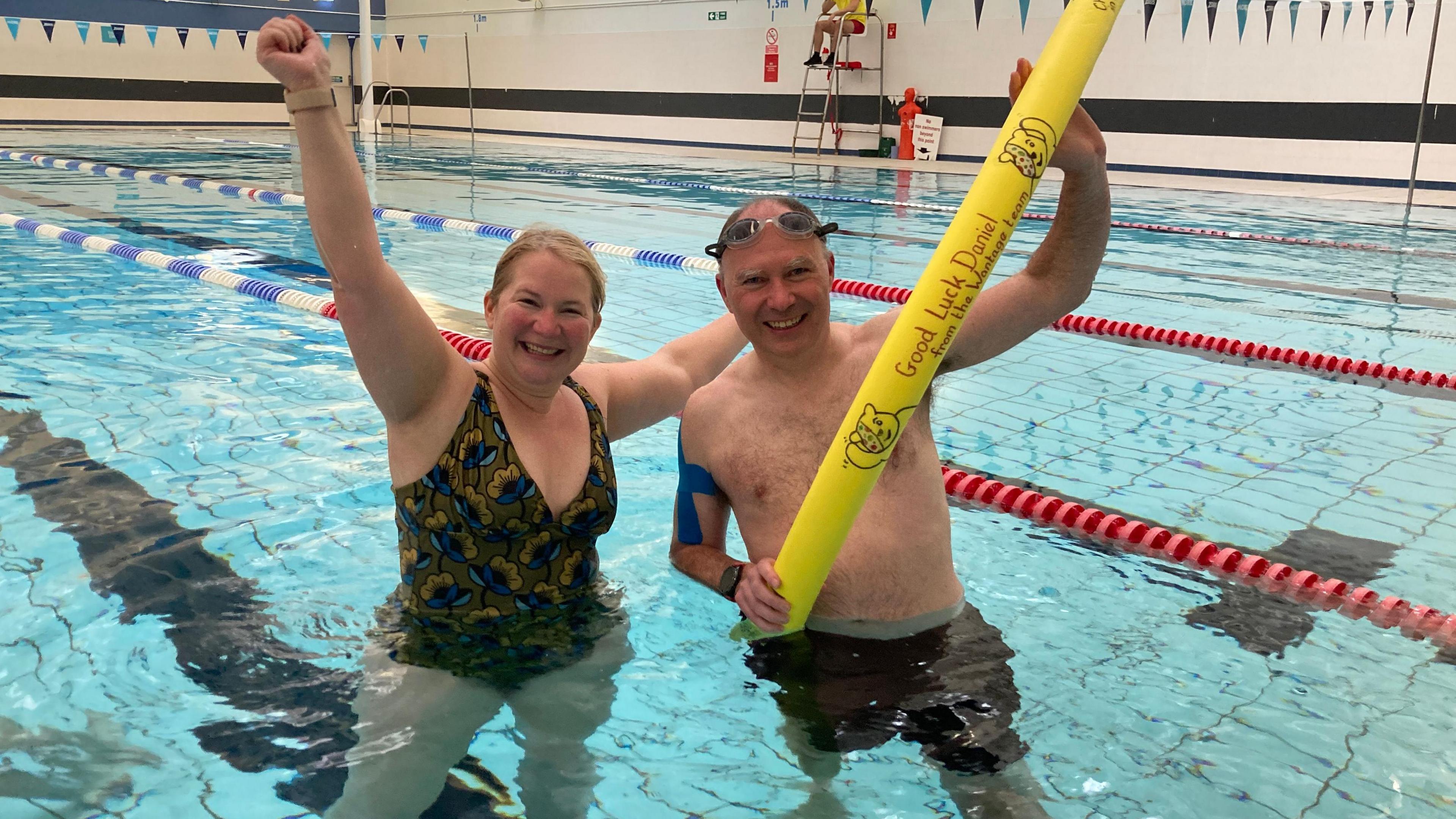 Sophie, wearing a one piece swimming costume with blue and yellow flowers on it, smiles and holds her arms aloft triumphantly. Dan, also smiling, is in dark swimming shorts and holds a long thin yellow swimming float that says 'Good Luck Daniel' on it. Both are standing in the pool.