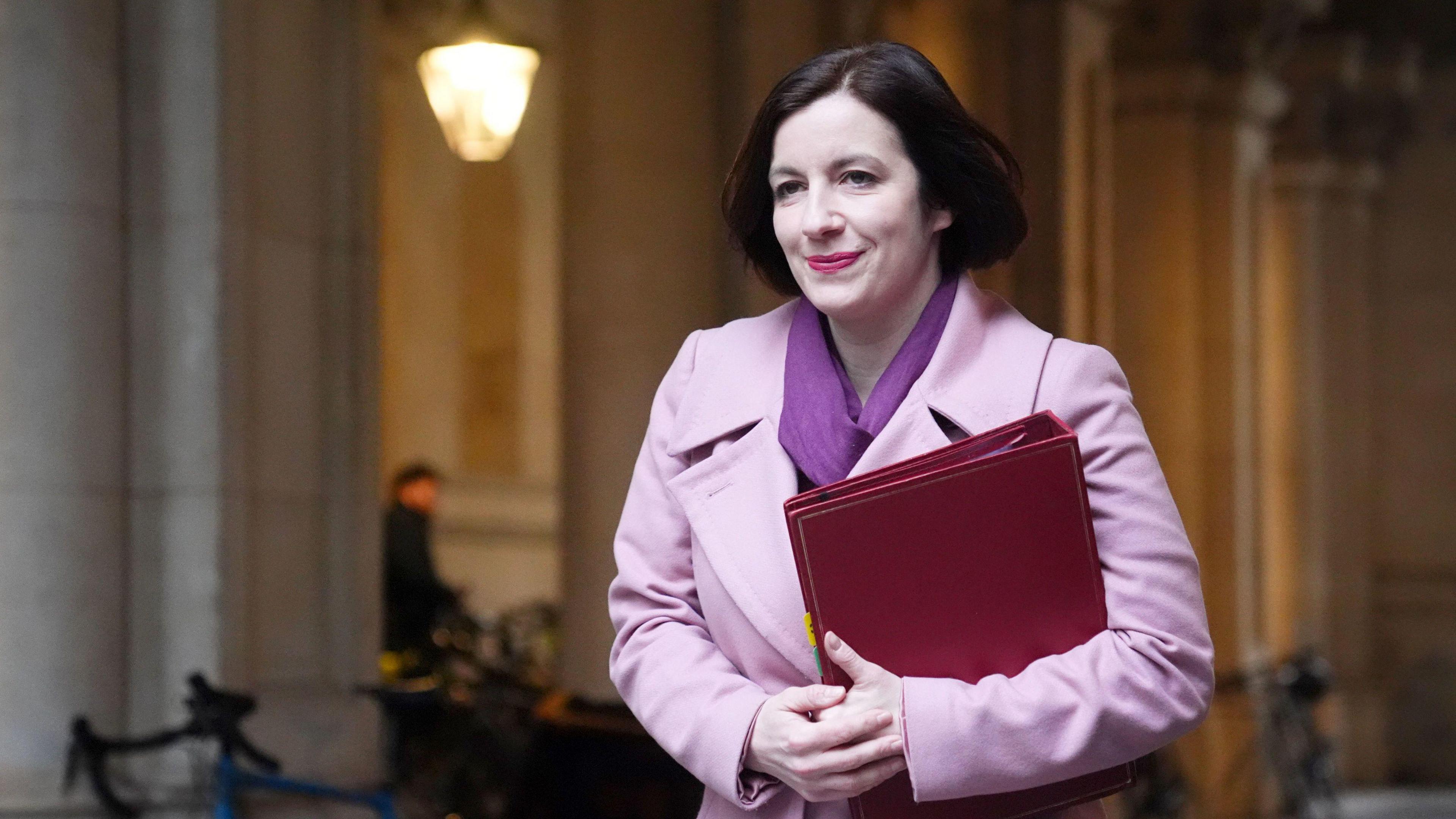 Bridget Phillipson walks along a street holding a red folder. She has long black hair and is wearing a purple scarf and a pink coat.