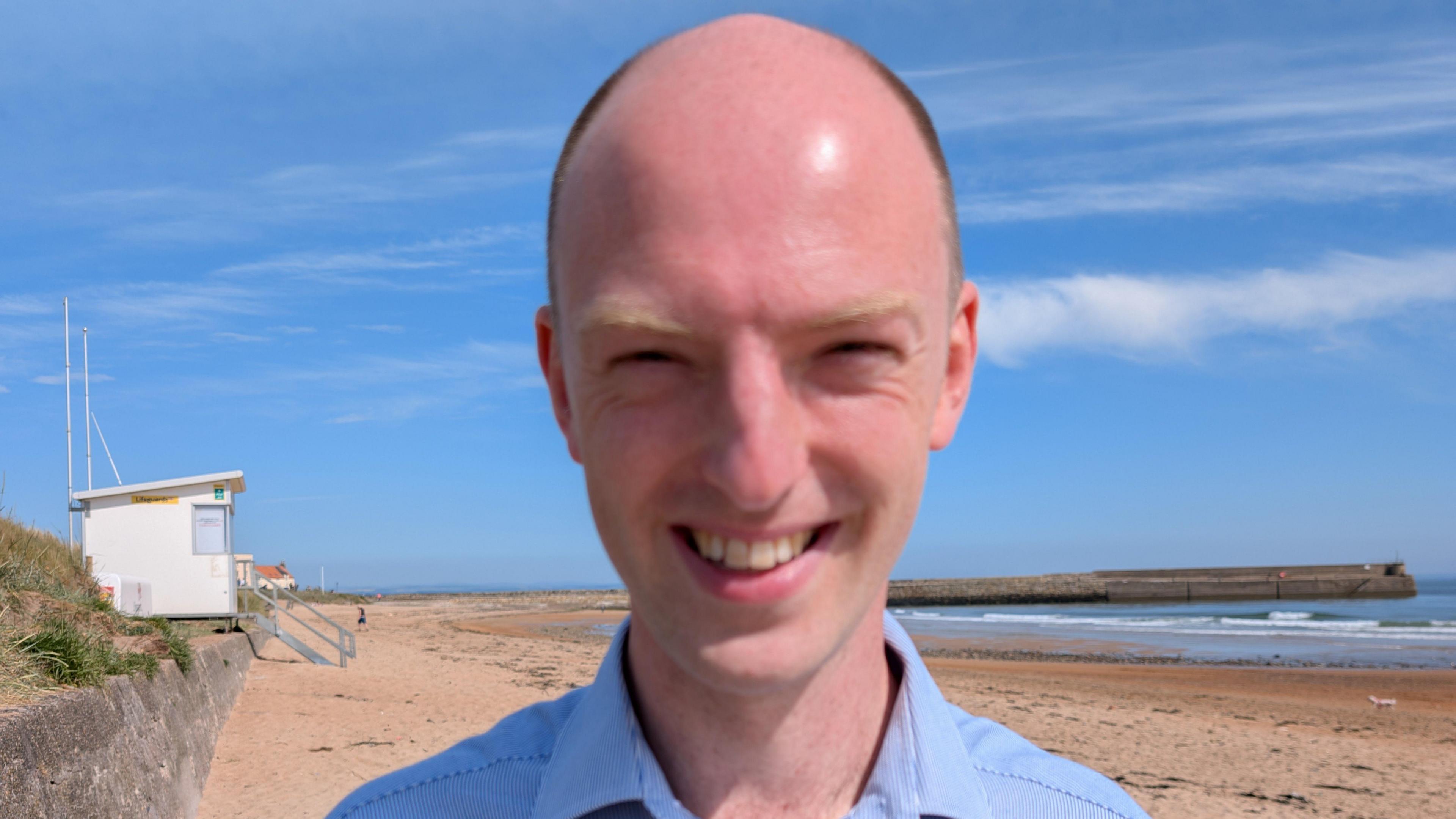 Dr Matt Patterson in a blue pin-striped shirt, standing on the East Sands at St Andrews on a sunny day.
