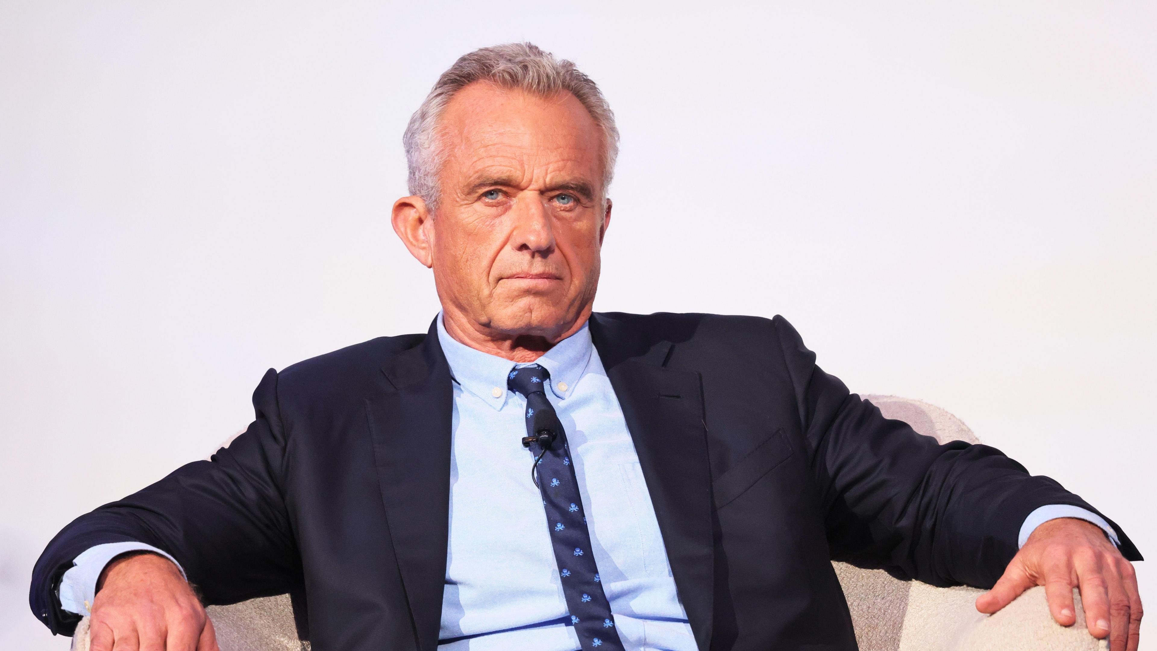 RFK Jr, wearing a suit, sits in an arm chair with his arms on the sides, with a neutral expression