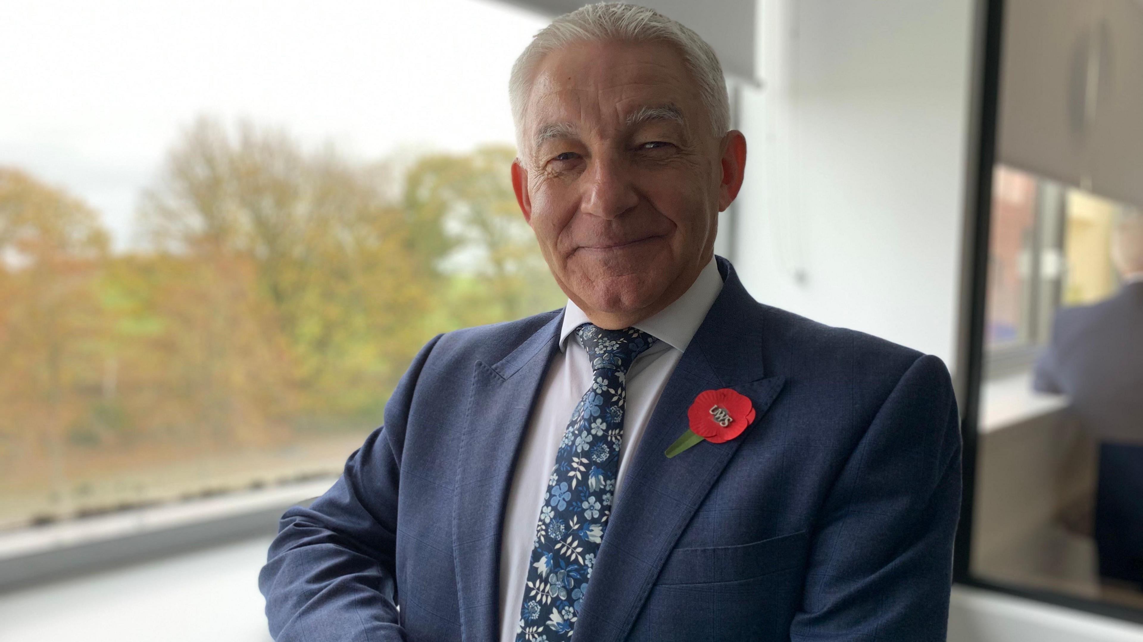 A man with grey hair in a blue suit with a poppy in the lapel and a white shirt and blue, floral-patterened tie