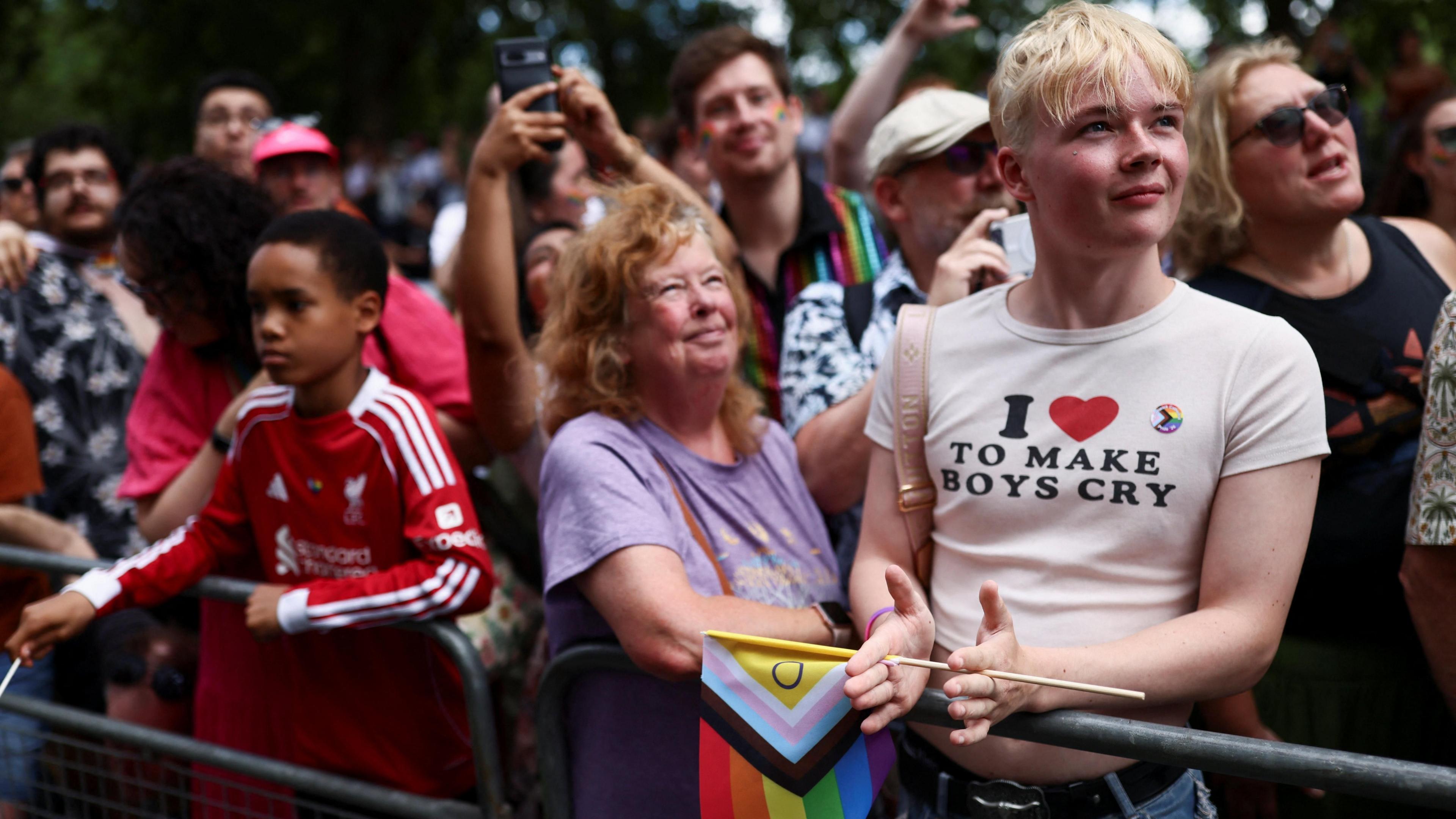 A crowd of spectators watching the parade from behind a metal barrier. In the foreground, a young person with short blonde hair claps while wearing a white cropped t-shirt that reads "I HEART TO MAKE BOYS CRY."