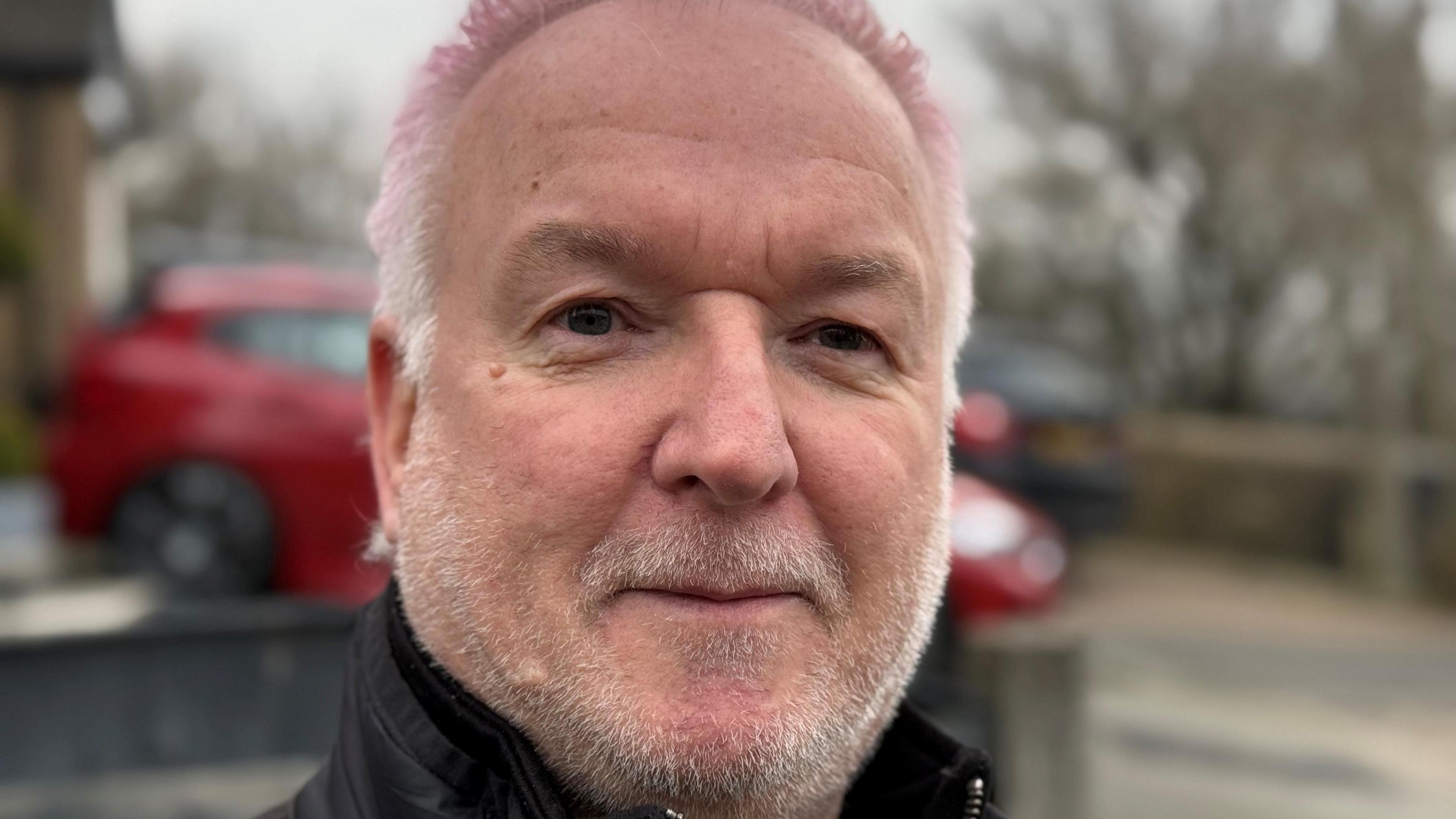 Gary Hough with cropped white hair tinged with a purple/pink dye wearing a black zip up jacket