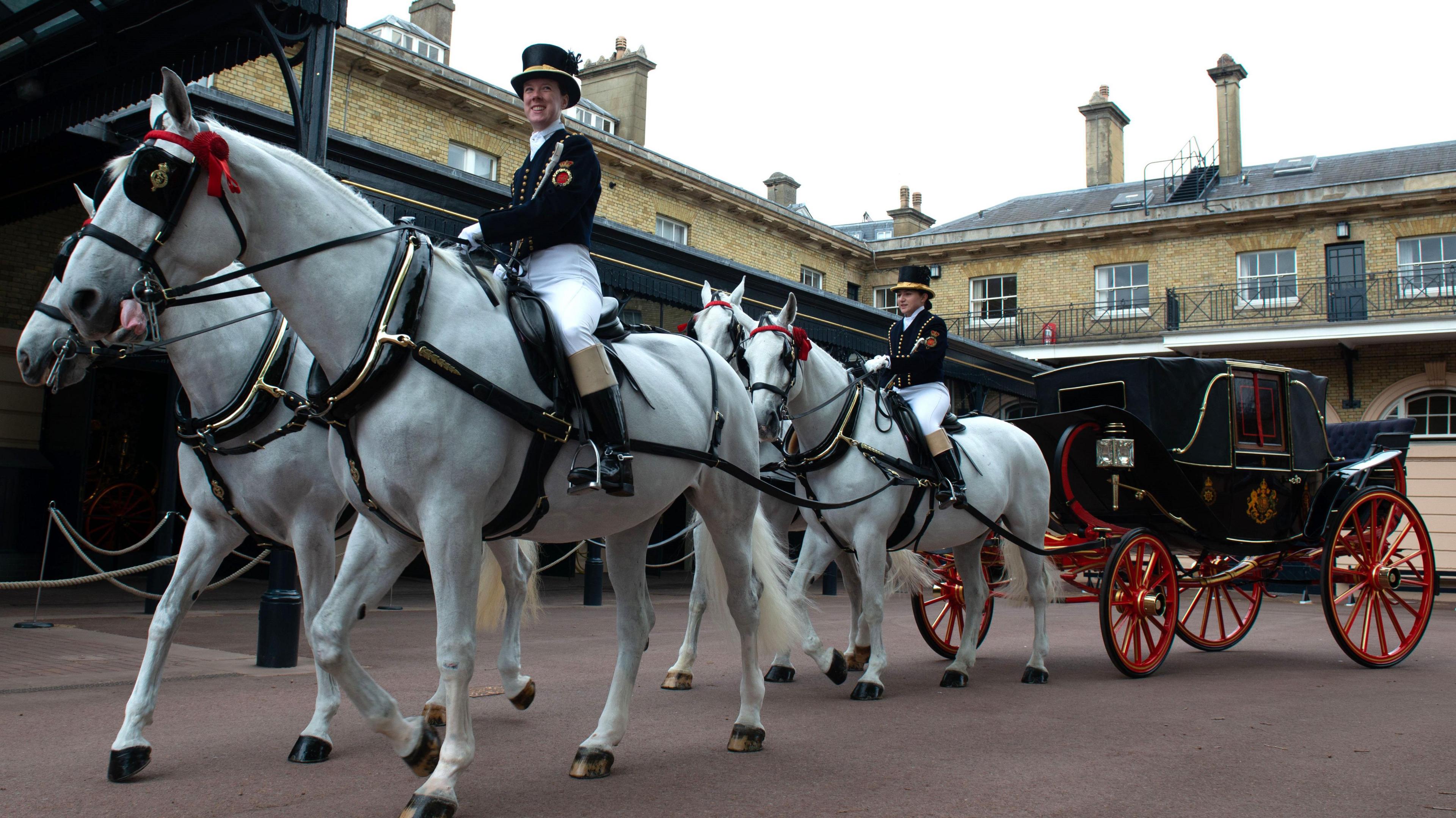 Four white horses pull a black carriage with red wheels past a 19th century two storey brick building. Two of the horses are mounted by grooms in black jacket and top hat and white breeches.
