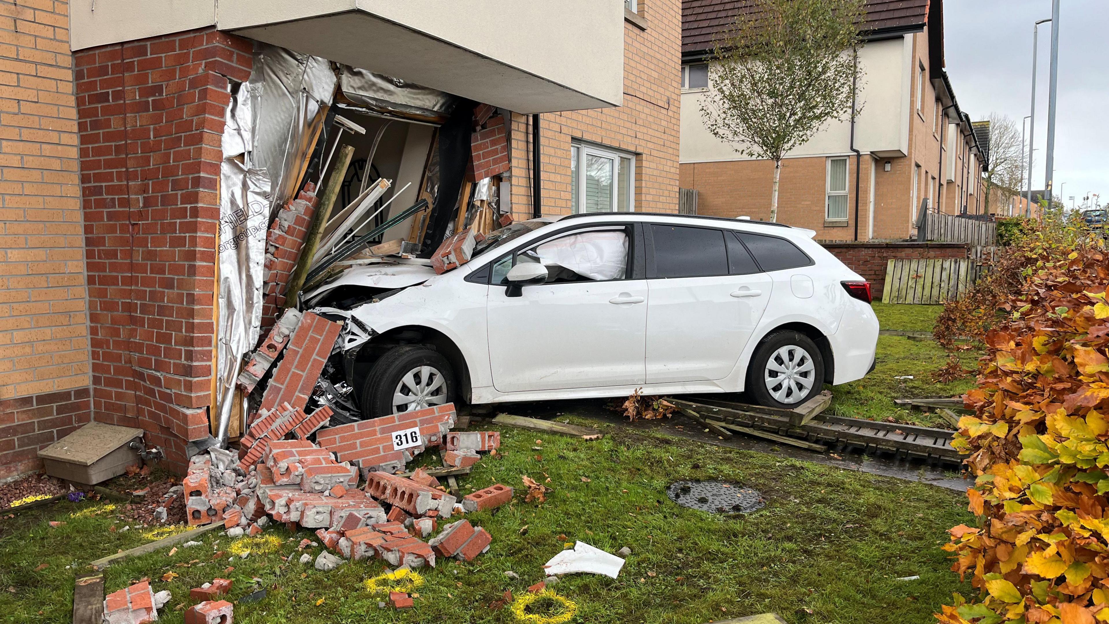 A white car that has crashed into the front of a house. There are bricks lying around the car and the front garden , and the car's inflated airbags can be seen through the windows.