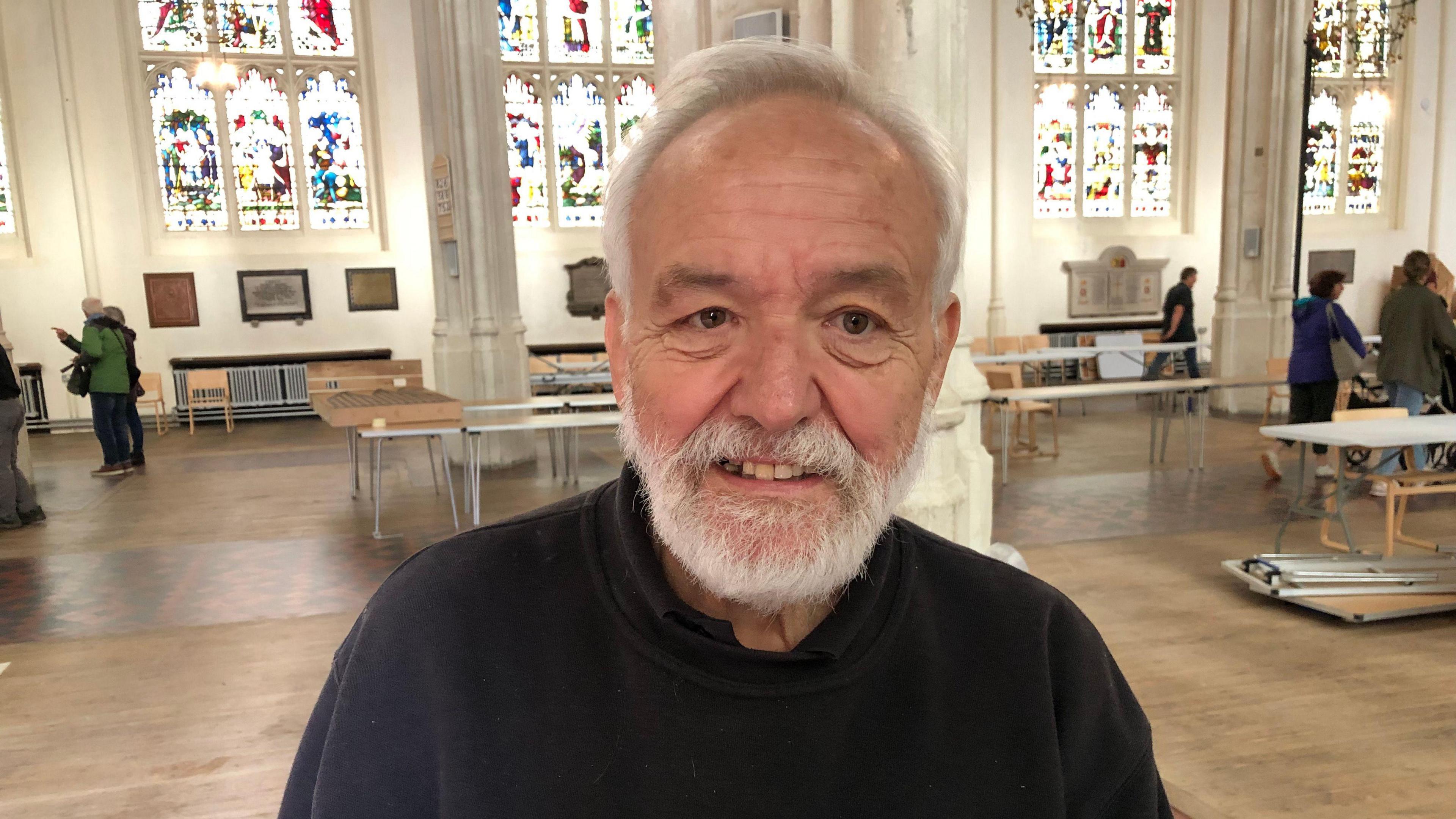 A photo of Chris Day inside the cathedral. He is smiling to camera. He has grey hair and a grey beard. He is wearing a black jumper with a black polo top underneath. Pillars and stained glass windows are behind him. People are pictured behind him as well as tables that will hold the model railway tracks.