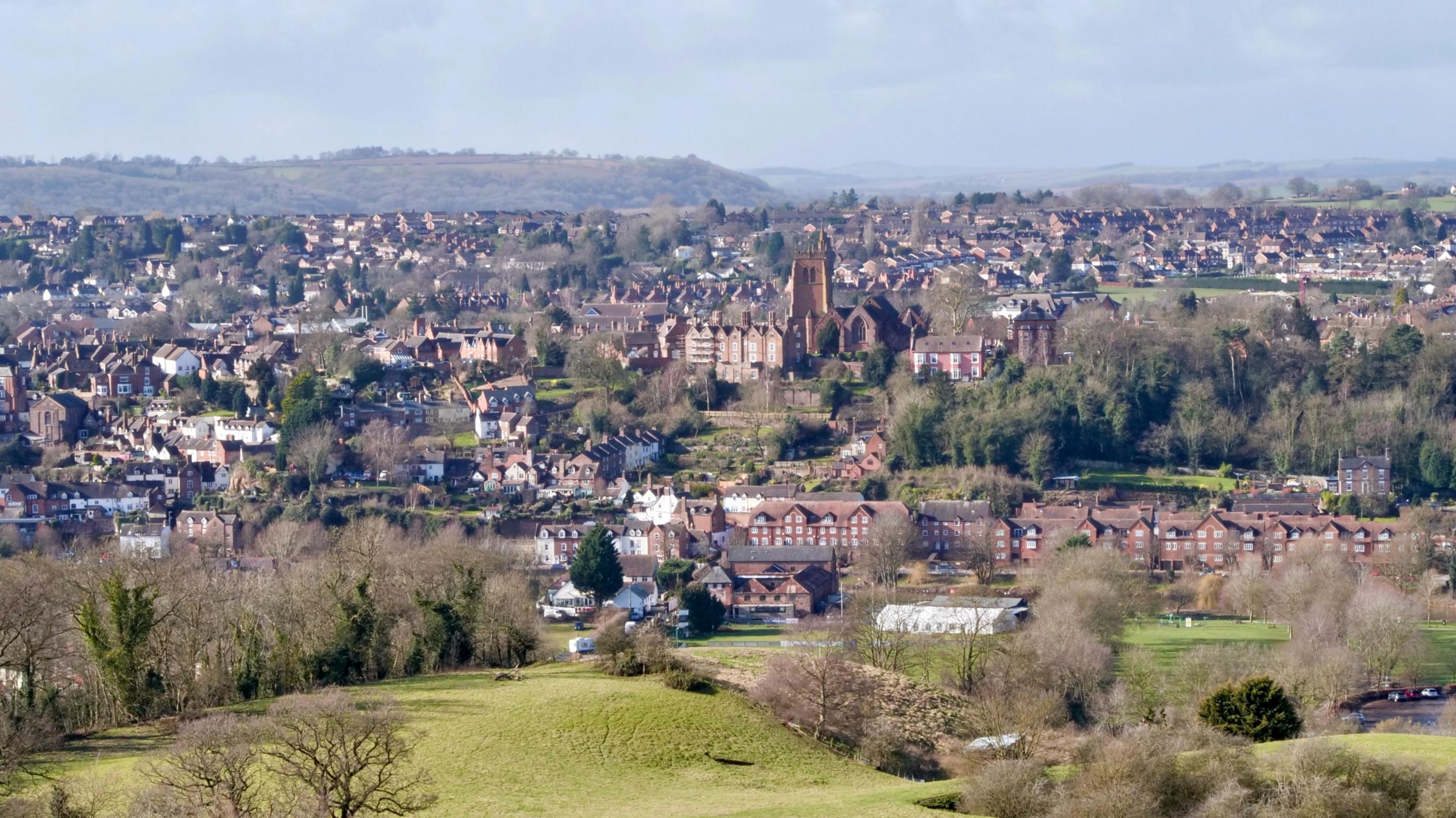Bridgnorth photographed from a hill overlooking the town. In the foreground are green fields before a line of brown riverside homes and then up to the town with the skyline dominated by a sandstone church