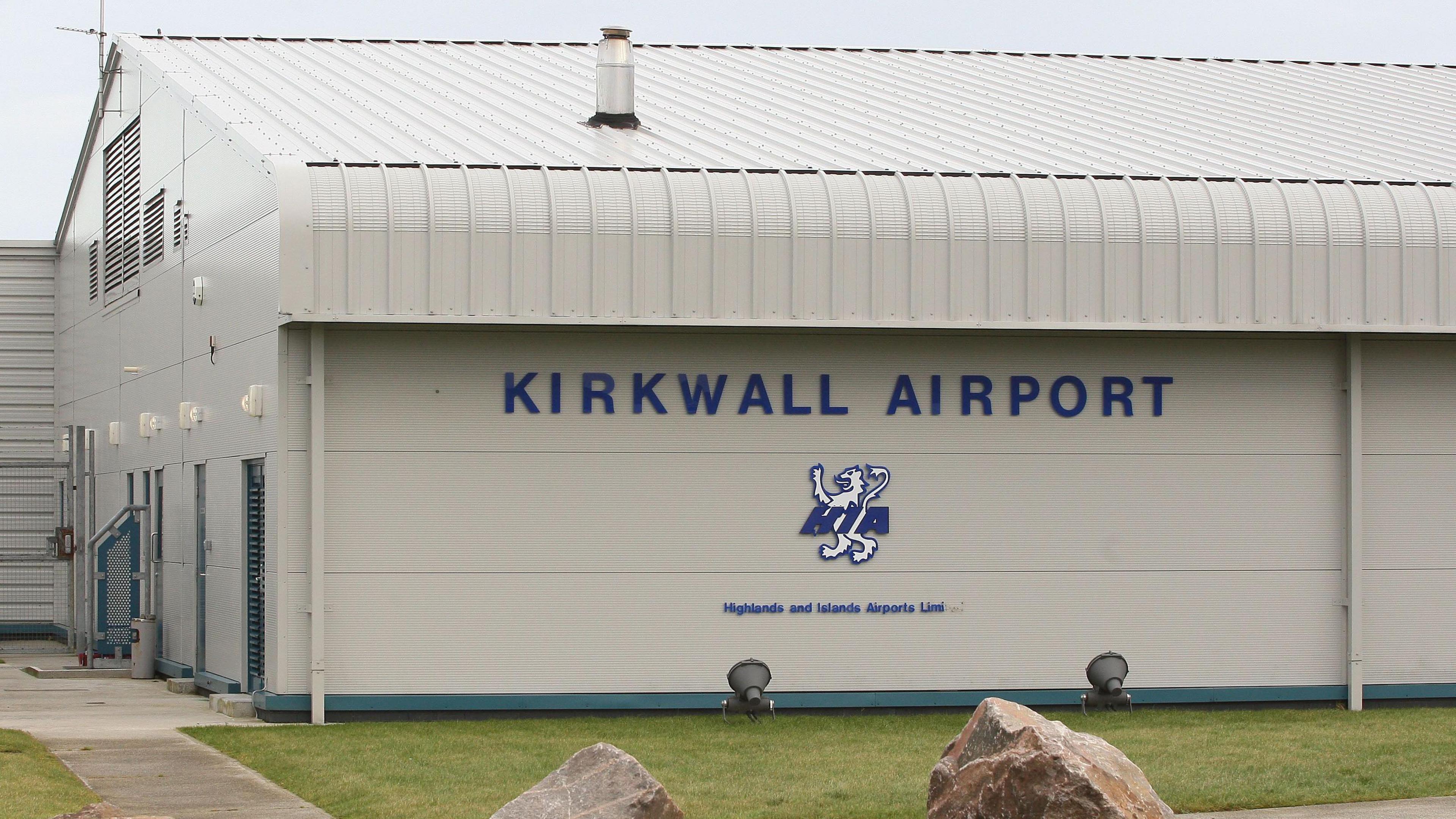 A general view of the Kirkwall Airport terminal building. It is grey with the name of the airport and a lion motif in blue. There is green grass in the foreground and the sky above is grey.