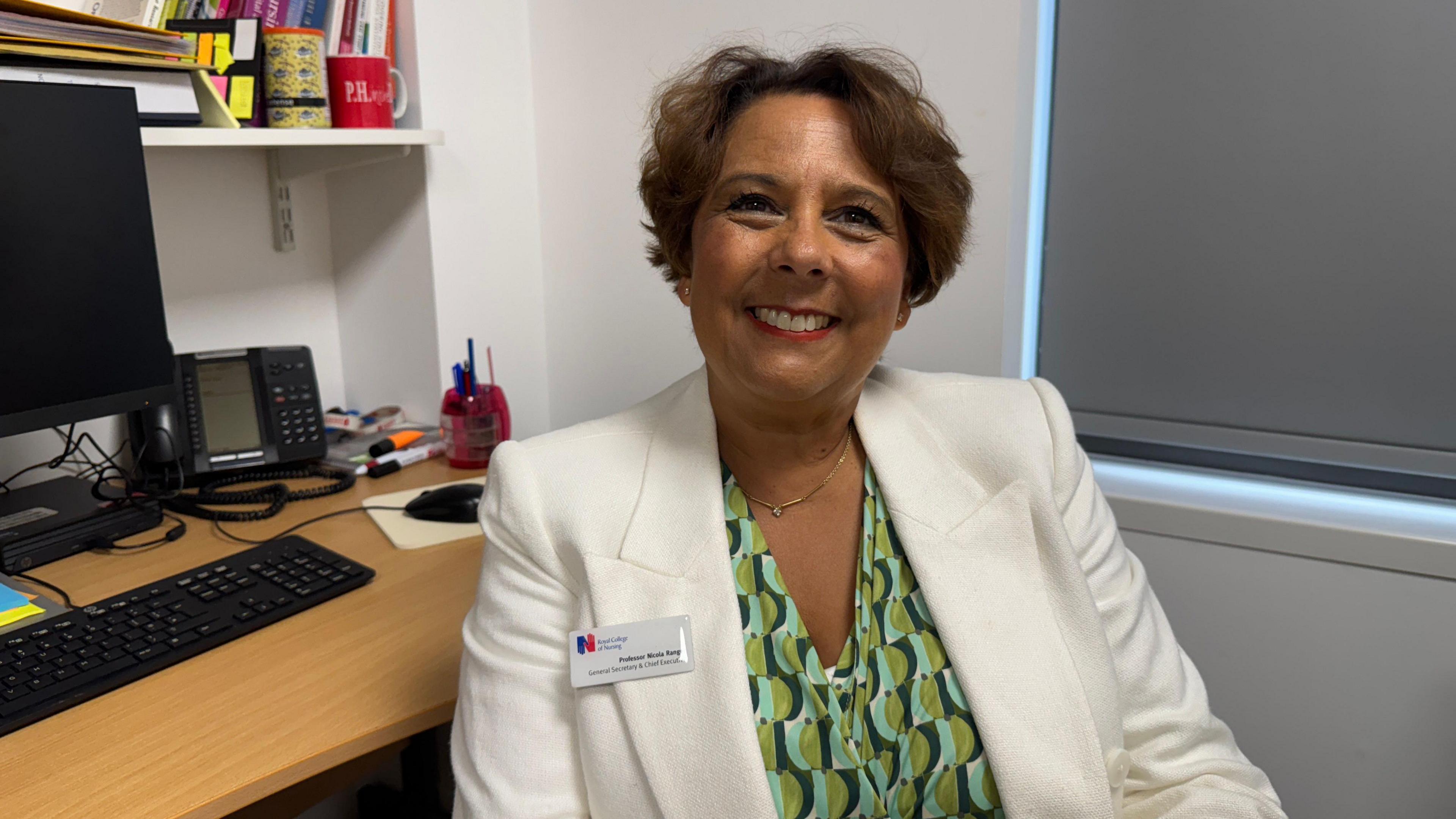 A woman with short brown hair smiling wearing a white blazer and a patterned light green, dark green and aqua blue top underneath. She is sat at a work desk with blinds closed behind.