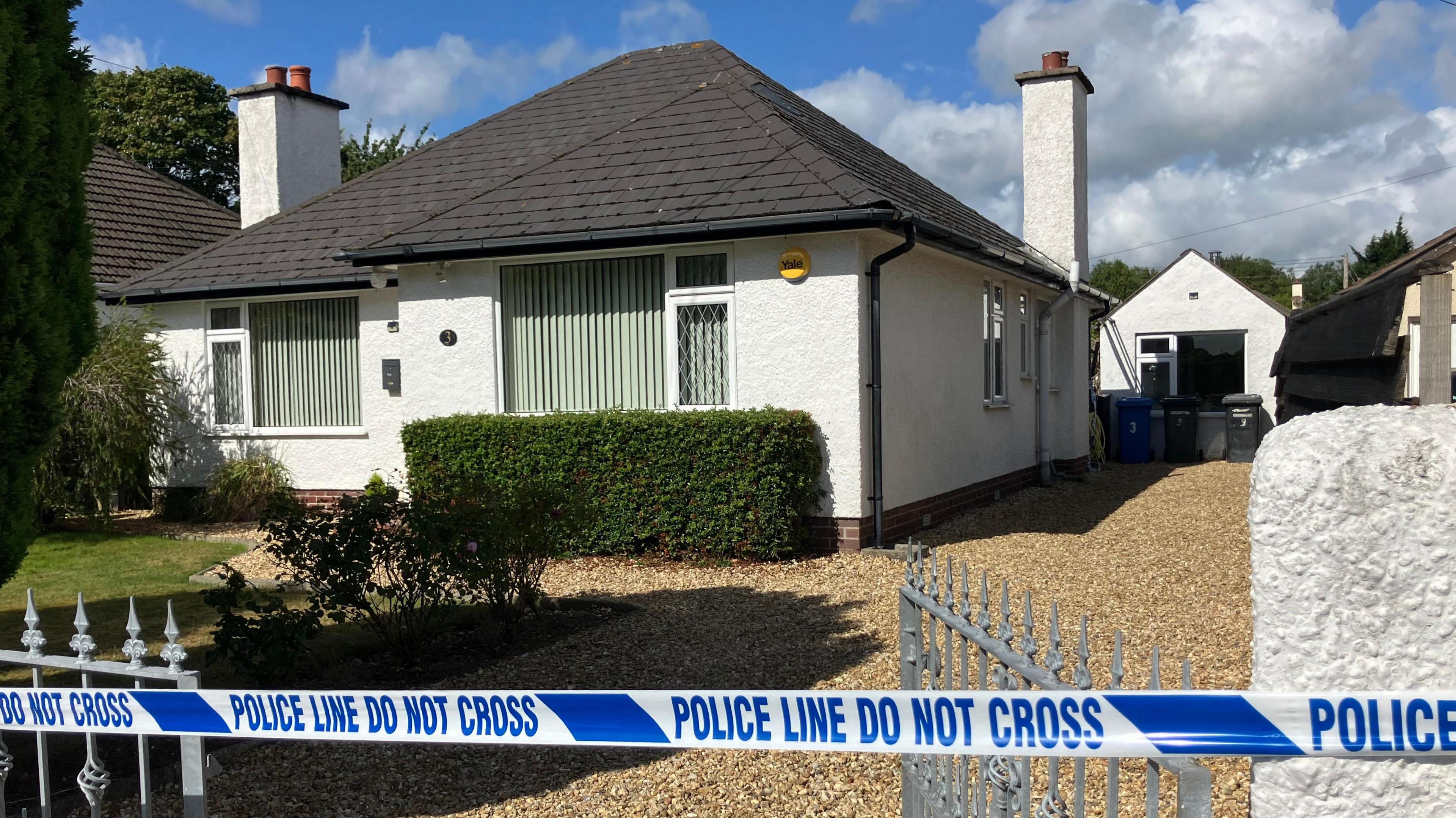 A white bungalow with a black roof with a garden in front of it and a stoned driveway is cordoned off with blue police tape.