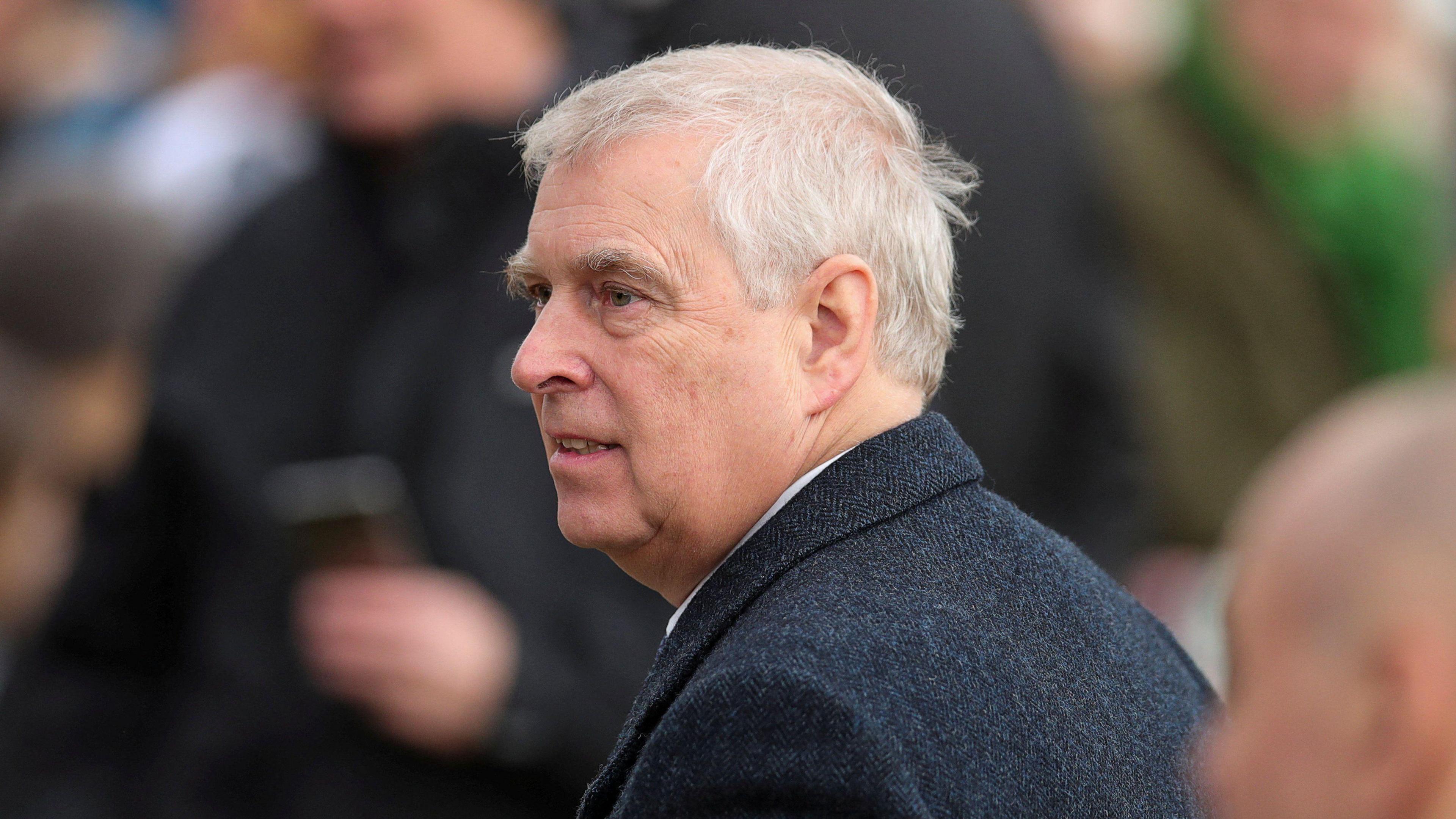 Prince Andrew wearing a dark overcoat over a white shirt, standing outdoors among a group of people. The background shows blurred figures