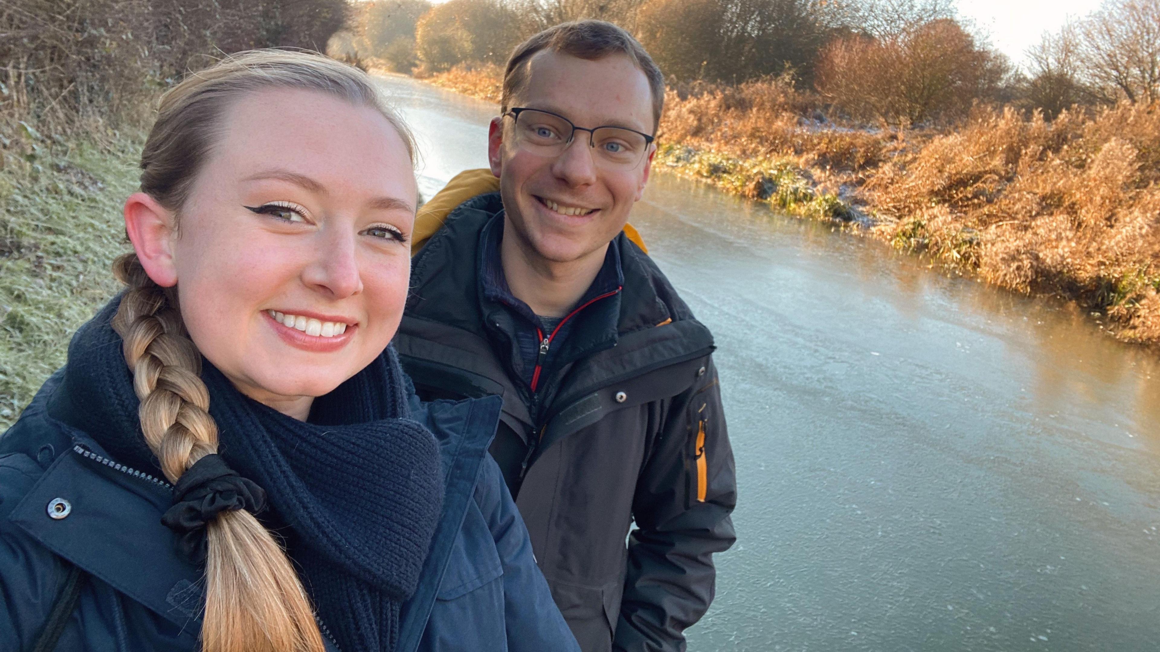 Alexandra Petri and Ross Jones wearing winter coats and standing beside a frozen river