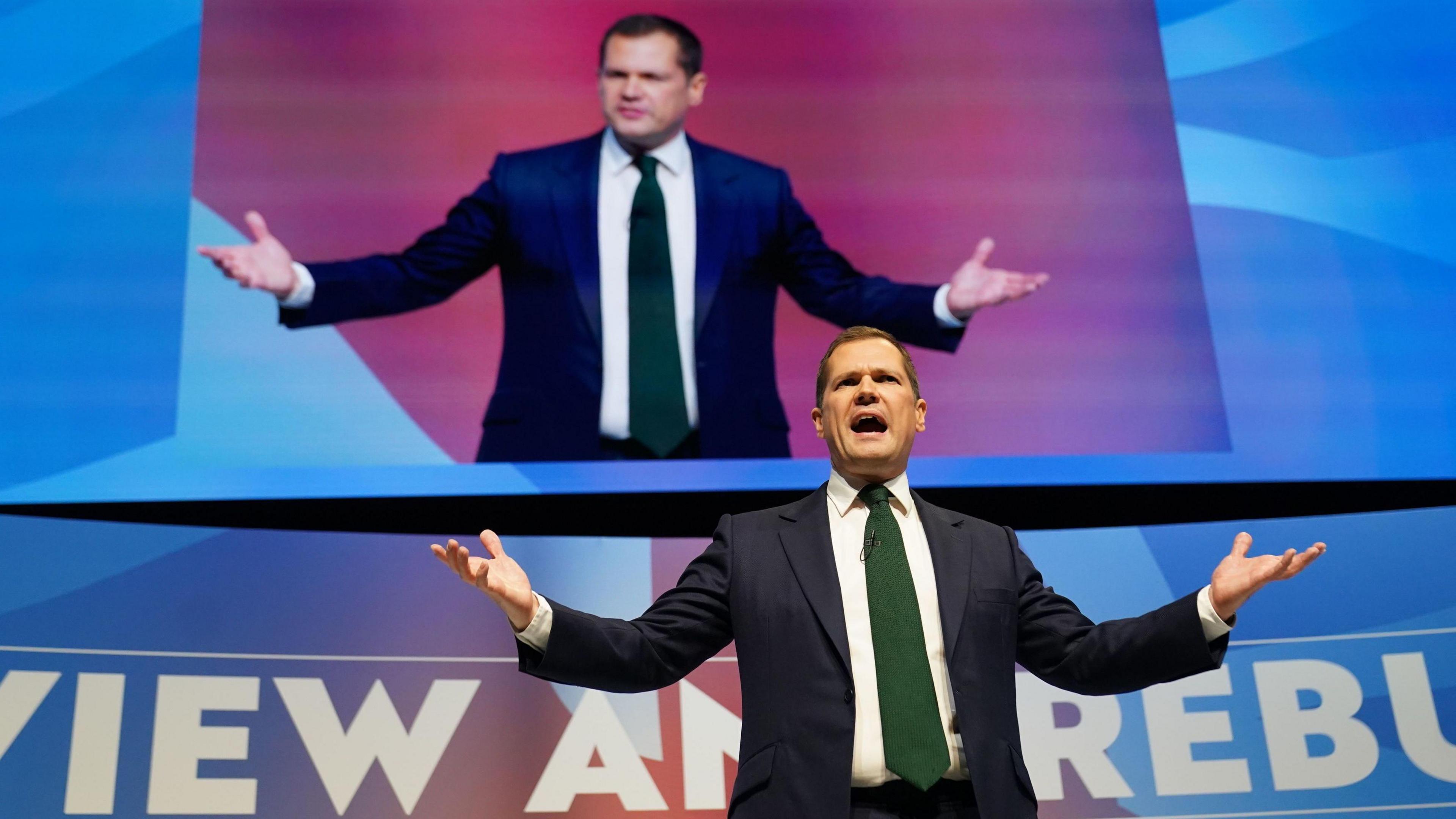 Robert Jenrick wearing a blue suit, delivering a speech on stage at the Conservative Party conference in Birmingham