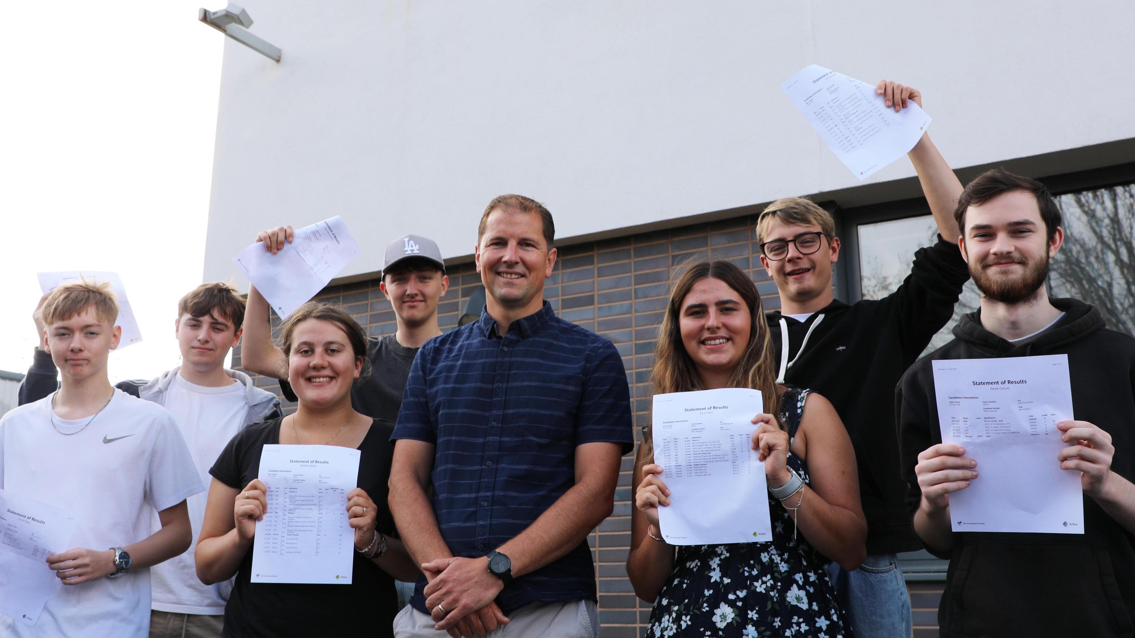 A group of Arnewood School students stand outside the school building with Mark Colman, head of sixth form. Everyone is smiling at the camera. There are three male students and one female student to the left of Mr Colman, holding pieces of paper with their results on either in front of their chest or in the air. Another two male students and a female student stand to the right of Mr Colman holding their results. Mr Colman has short, light brown hair, wears a short sleeved dark blue striped shirt and beige chino trousers. He has his hands crossed in front of him in a relaxed pose. 