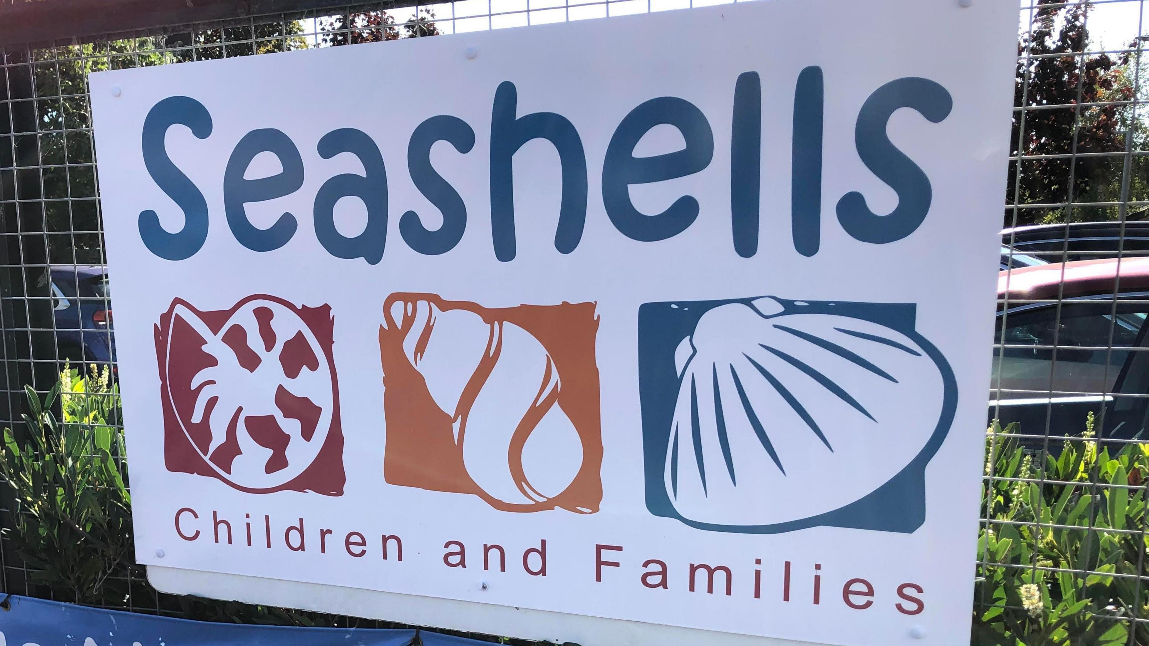 A white sign reads "Seashells children and families". It is on a fence with greenery and cars behind. 