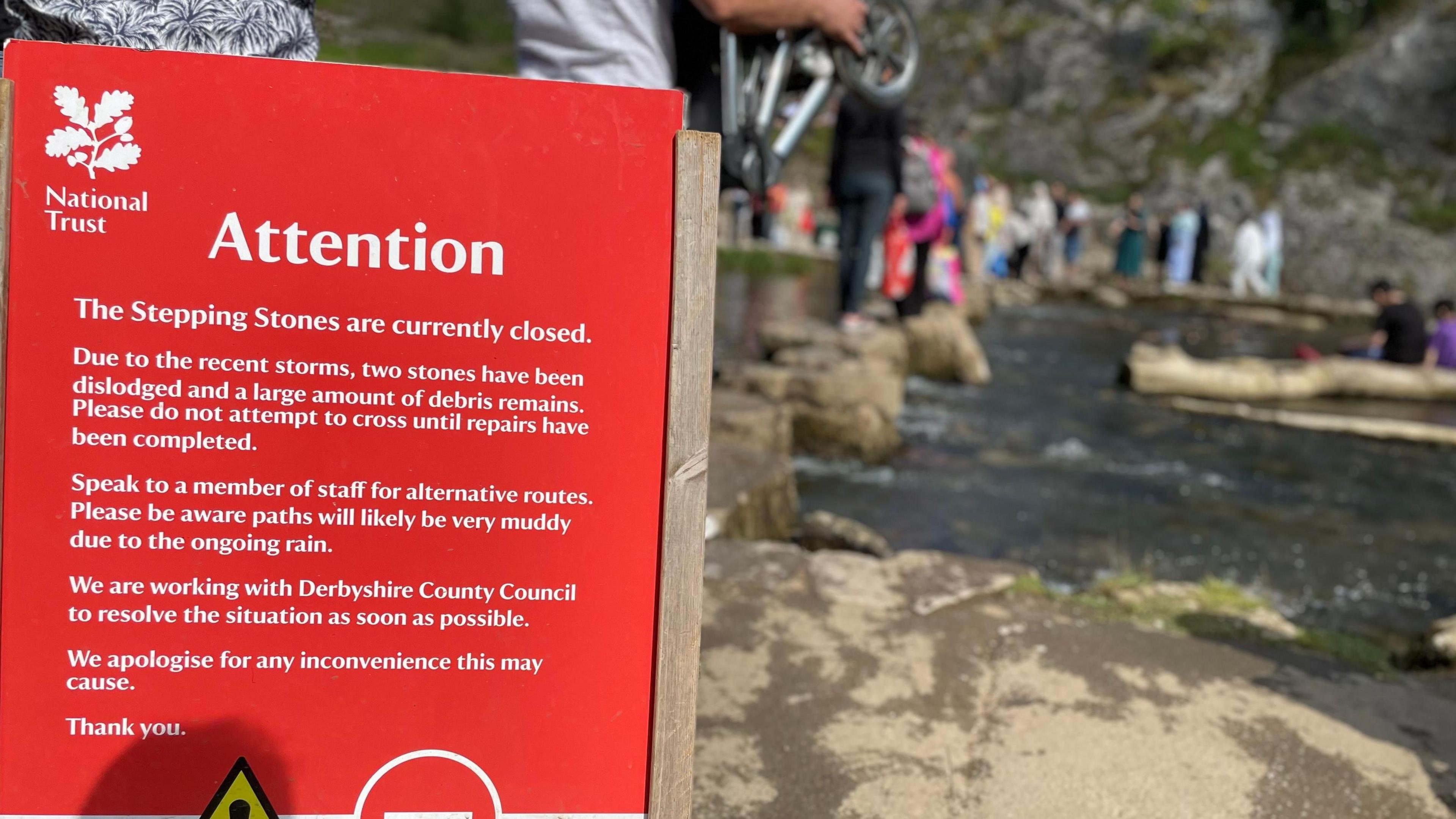 A sign advising the stepping stones are closed, with people crossing them in the background