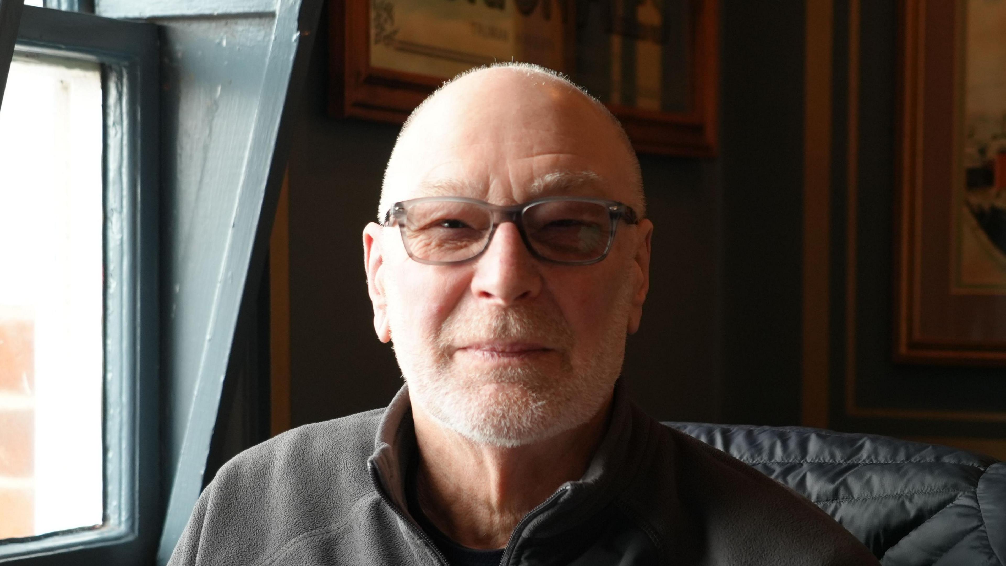 John Dupen, wearing glasses and a grey jumper, staring into the camera.