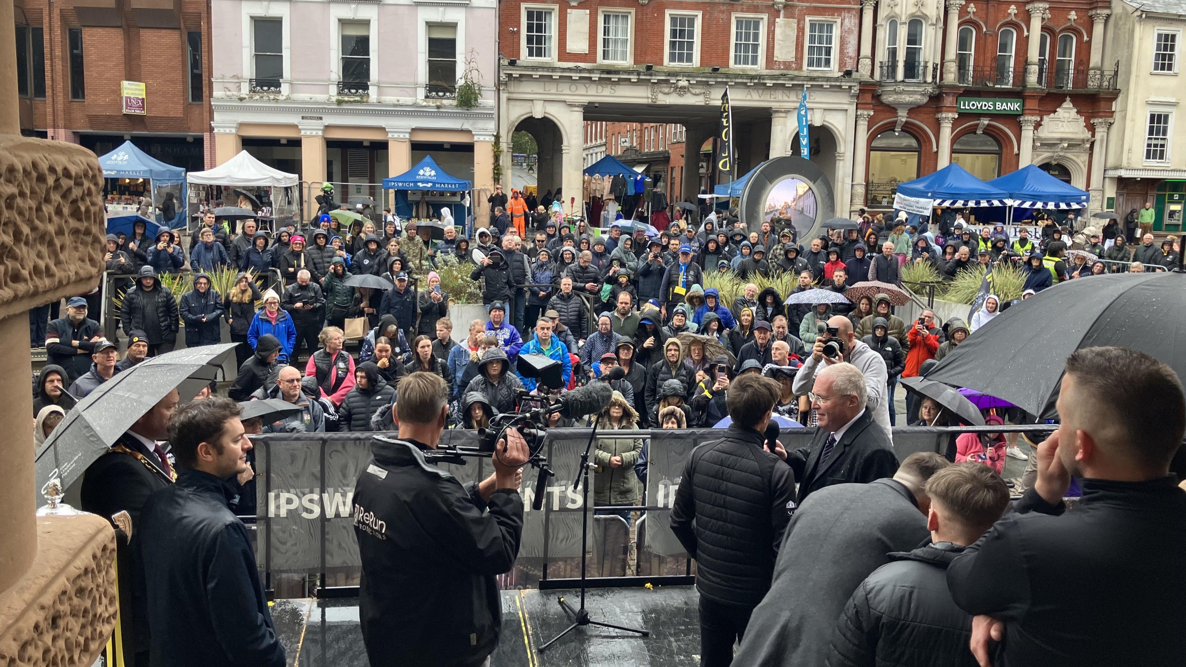 A crowd of people look toward the camera during a celebratory event at a town hall. They stand behind a fence watching as interviews are done on the steps. Some people with cameras can be seen. It is raining so many people wears coats and have umbrellas.