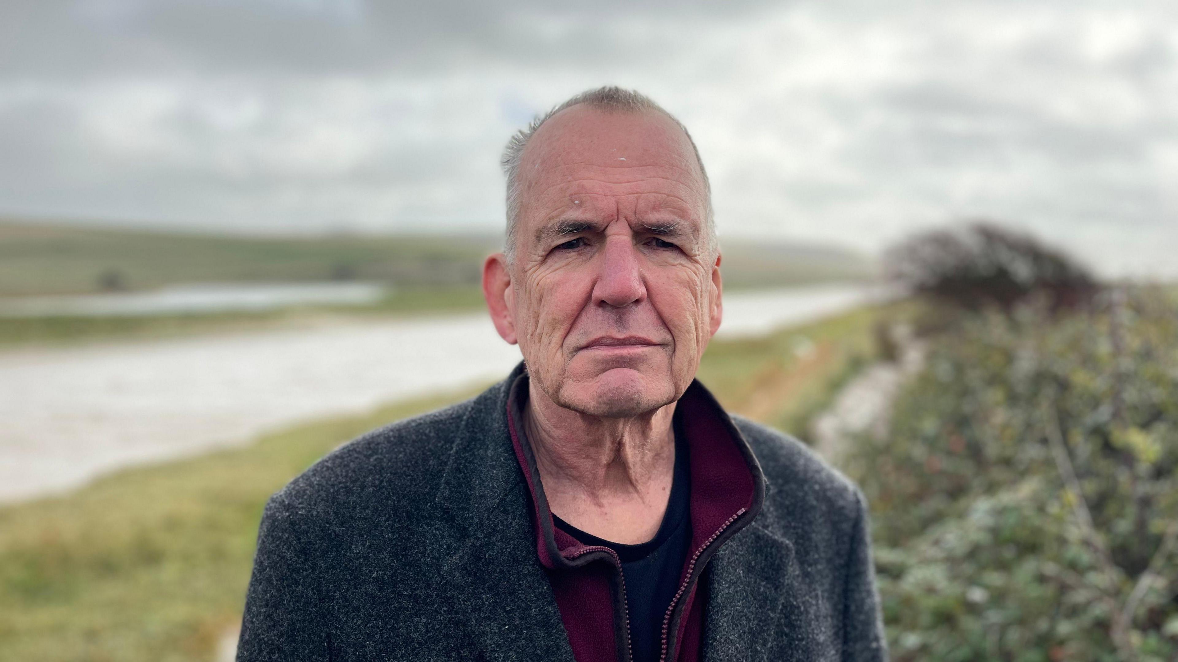 Chris Smith standing on the river bank which is due to be breached. He is wearing a wooly coat. The river is straight and has bushes on it