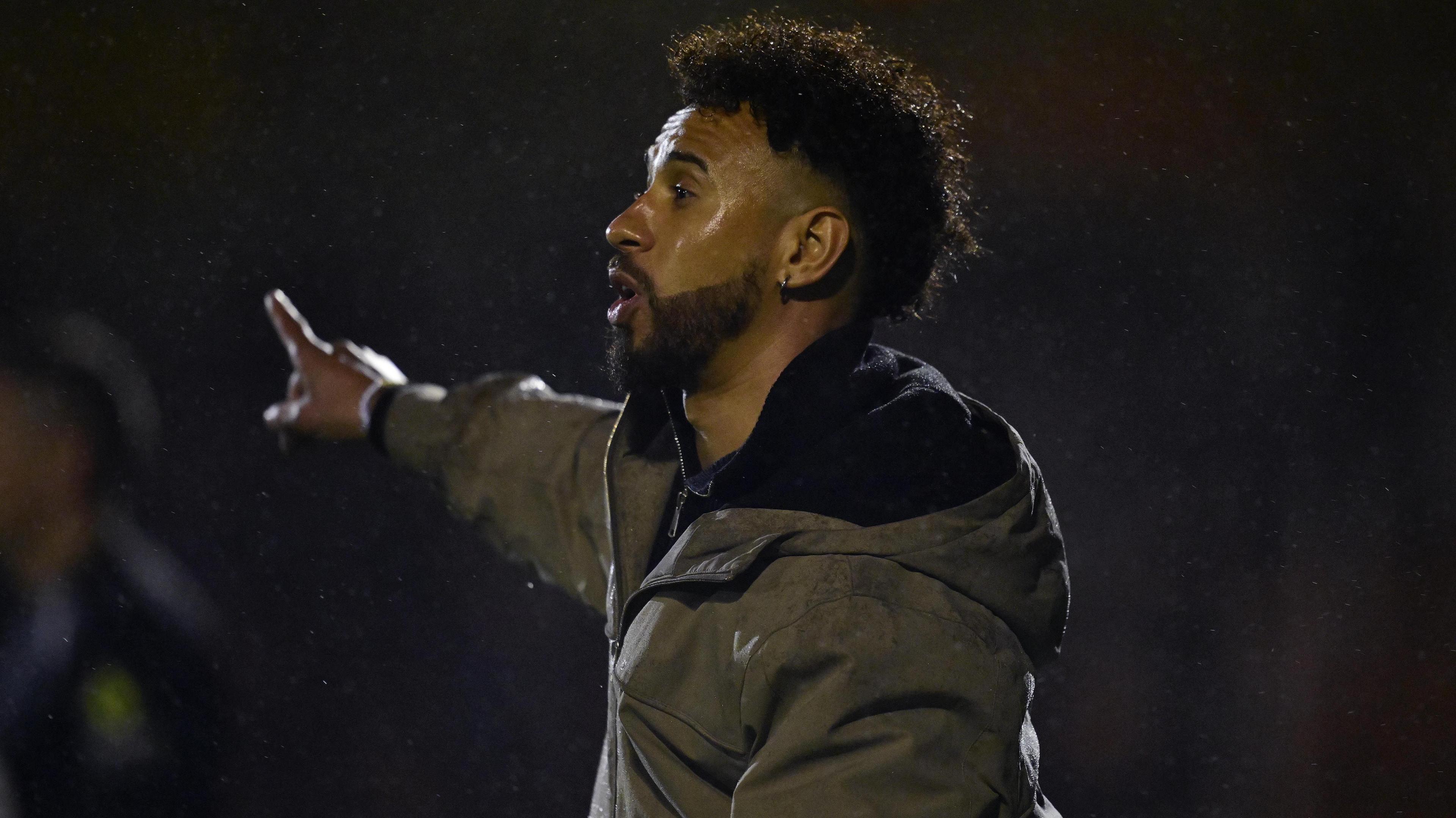 Sam Cox, wearing a green jacket, points while on the sidelines 