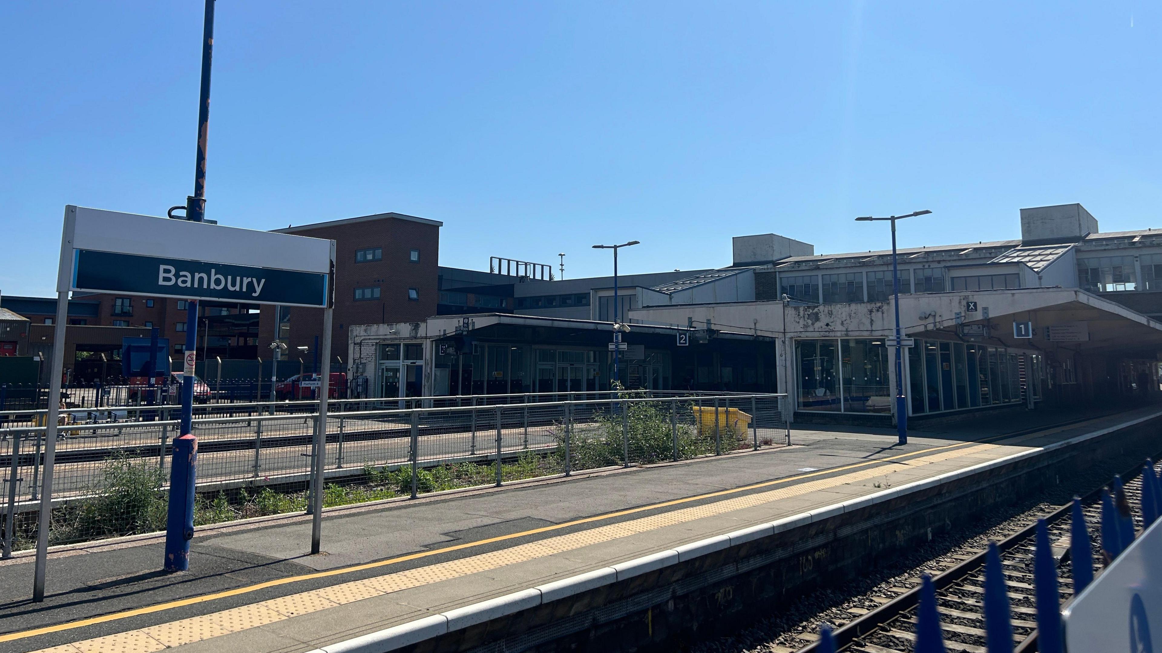 A sign saying Banbury sits above a train platform and railway line in the foreground, further platforms are visible in the distance. In the distance, an ageing building is also visible - this is the station itself