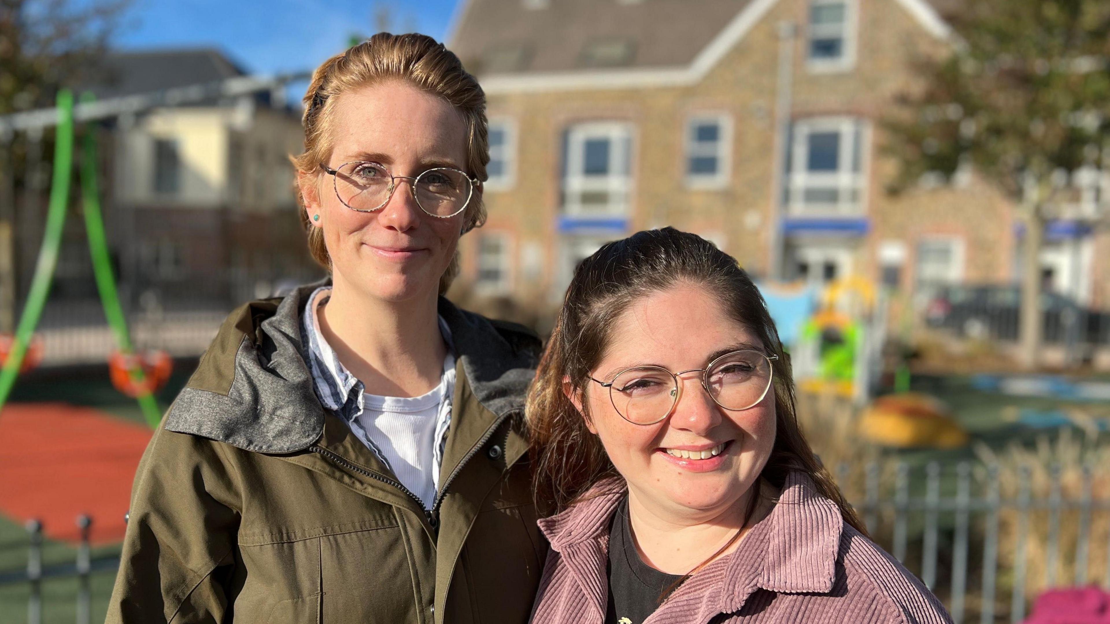 April and Lauren are standing close together in an outdoor playground. They are wearing jackets, and behind them is a play area with green swing frames and a soft red play surface. In the background, there are stone buildings with multiple windows and a clear blue sky.