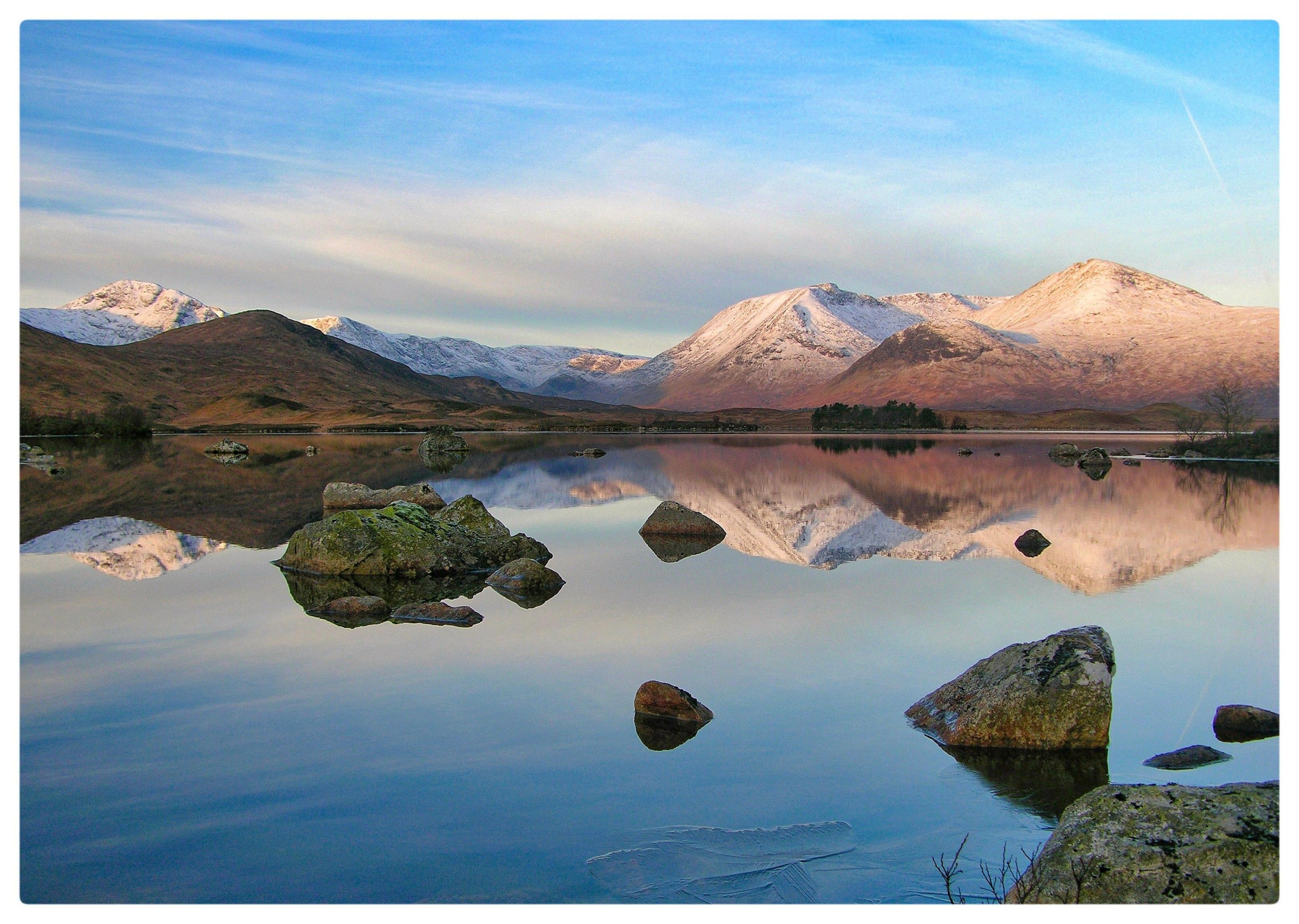 Snow-capped mountains rise under a soft blue sky, their peaks mirrored perfectly in a still, glass-like lake. Scattered rocks break the water’s surface, adding texture to the serene reflection.