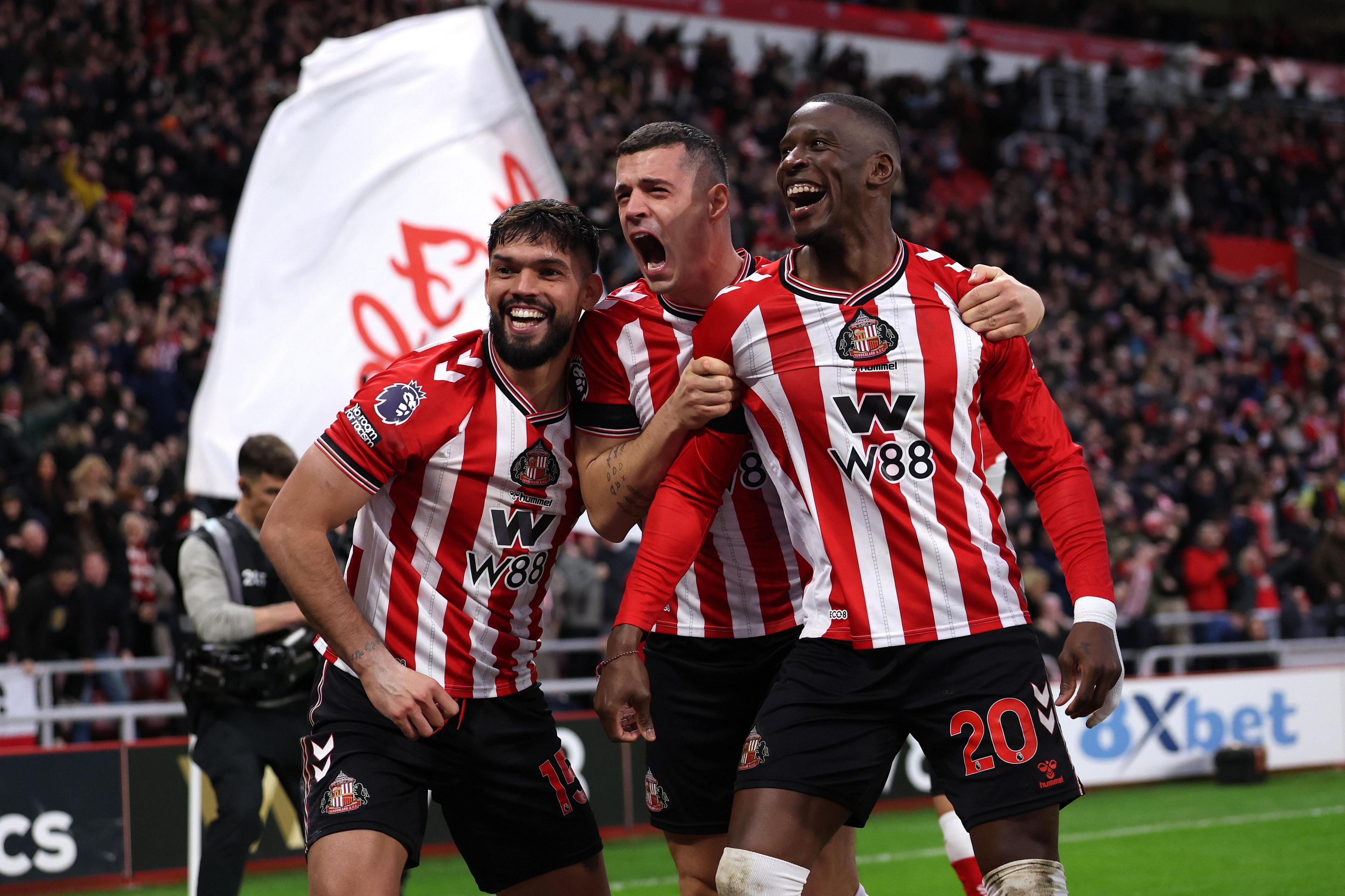 Tres jugadores del Sunderland que visten camisetas a rayas rojas y blancas celebran juntos frente a una gran multitud, con una bandera gigante del equipo ondeando detrás de ellos al margen.