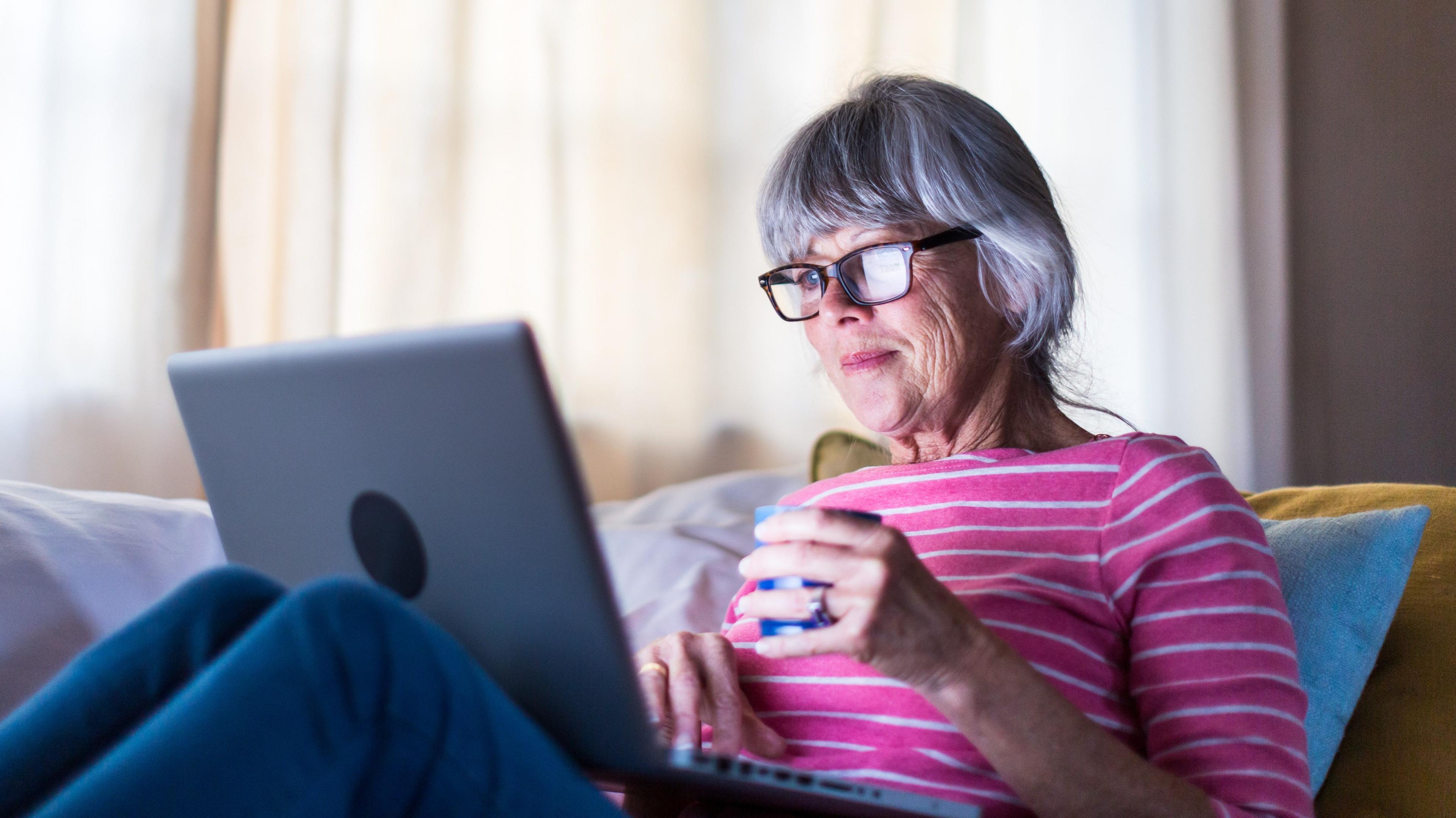 A woman with grey hair and glasses holds a mug in her hand as she looks at her computer while sitting on a sofa.