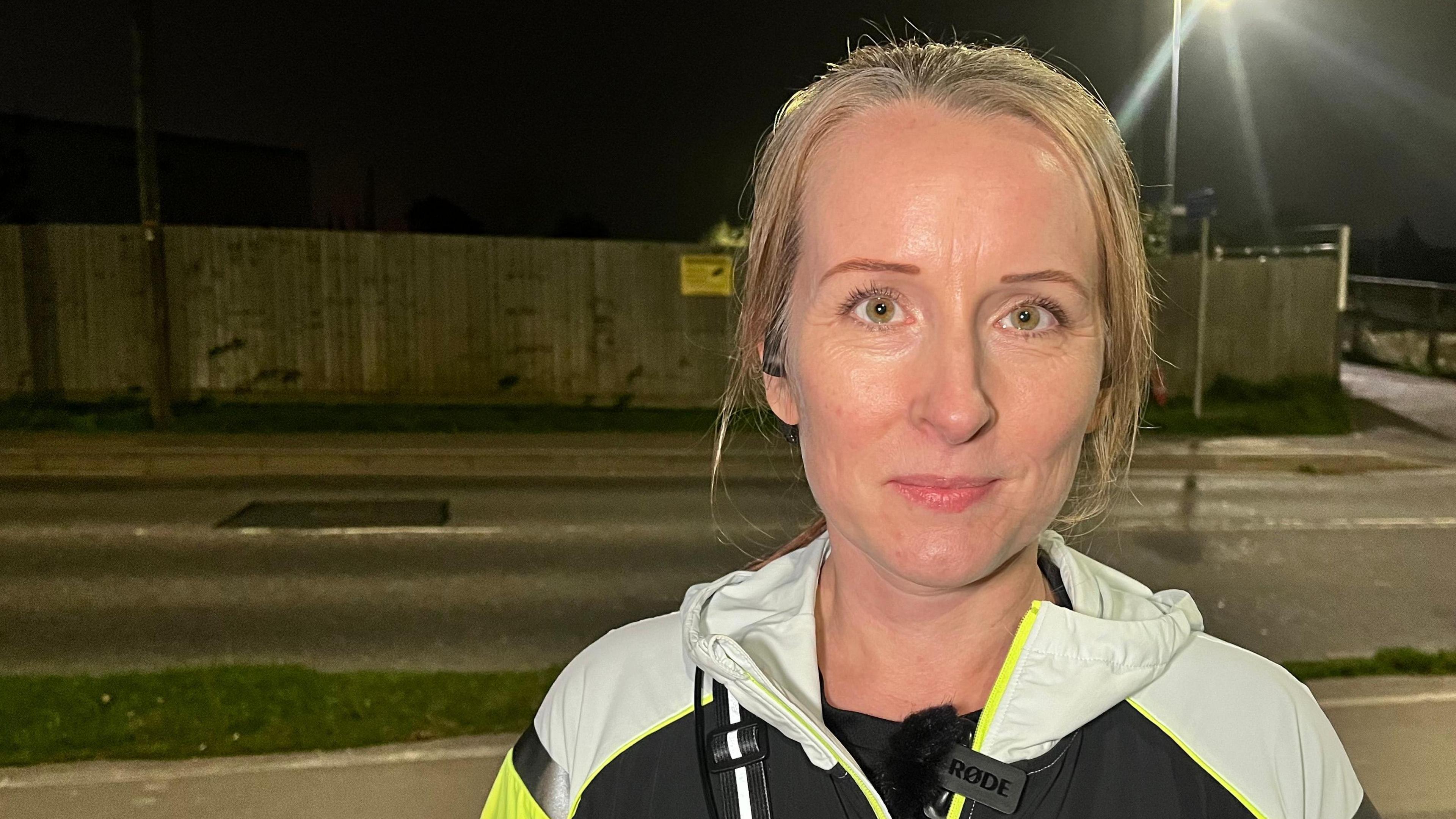 Melissa Hawkes stands in a street under a night sky. She has blond hair, tied back, and wears a black, grey and yellow running top.