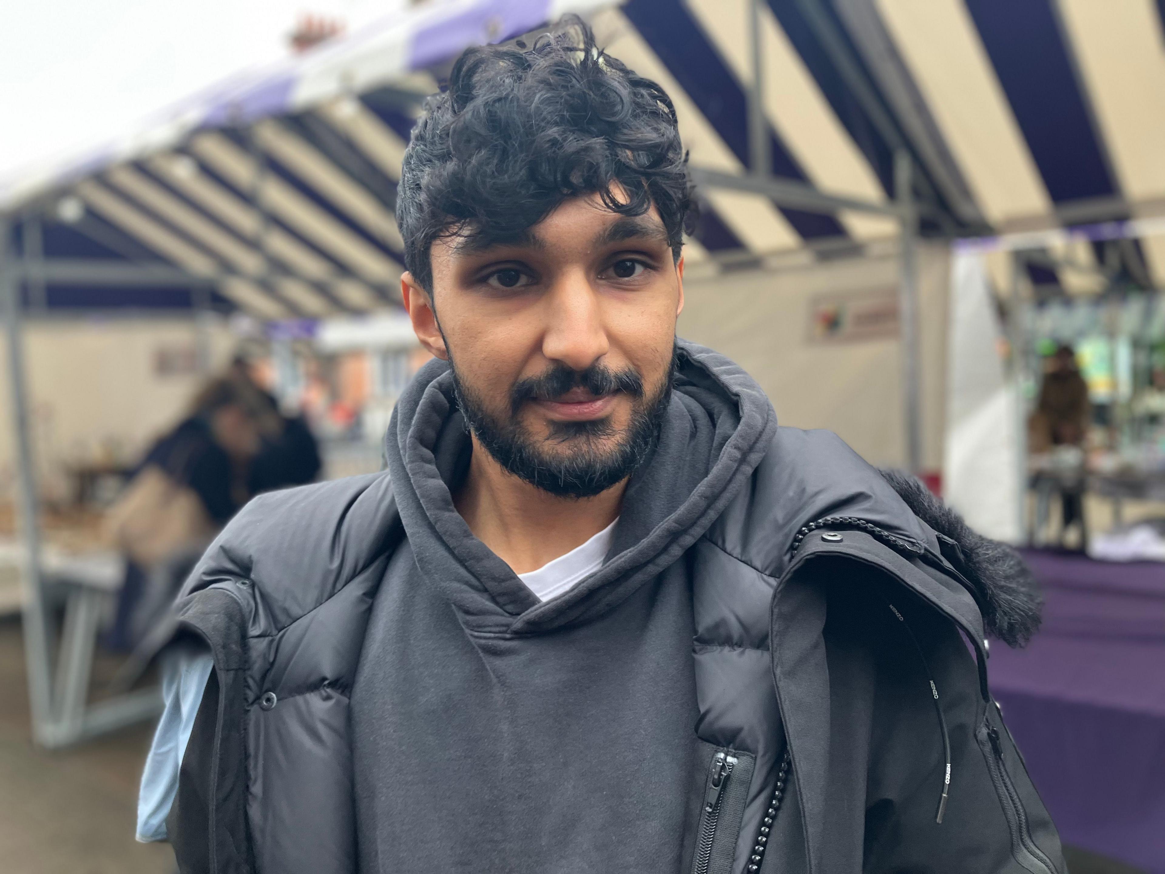 A young bearded man with unkempt hair dressed in a big black coat and a black hooded top stood in front of market stalls