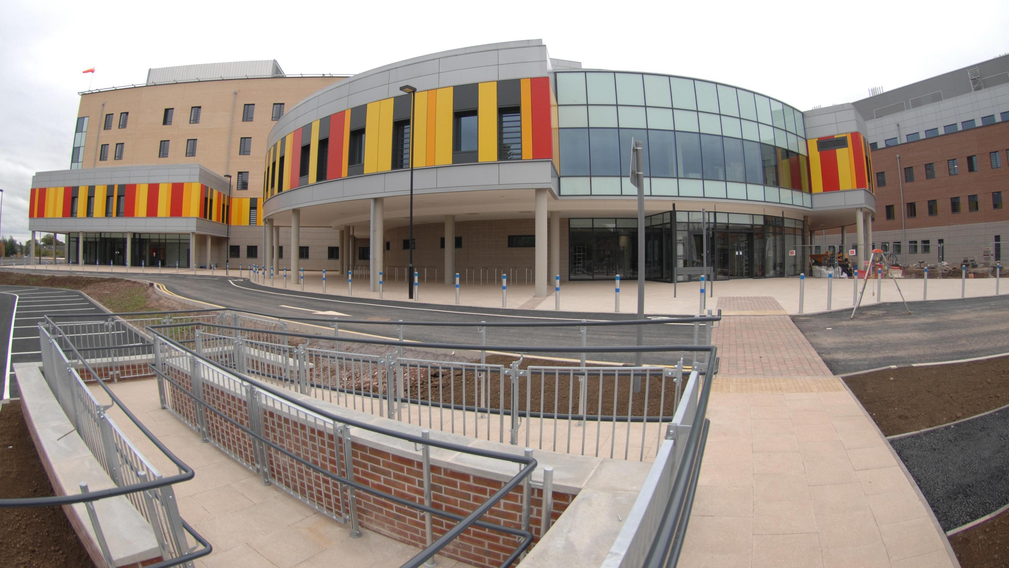 General view of Royal Stoke University Hospital. It is a large building with a semi-circular frontage with yellow, red and orange panels. There is a walkway in front of it.