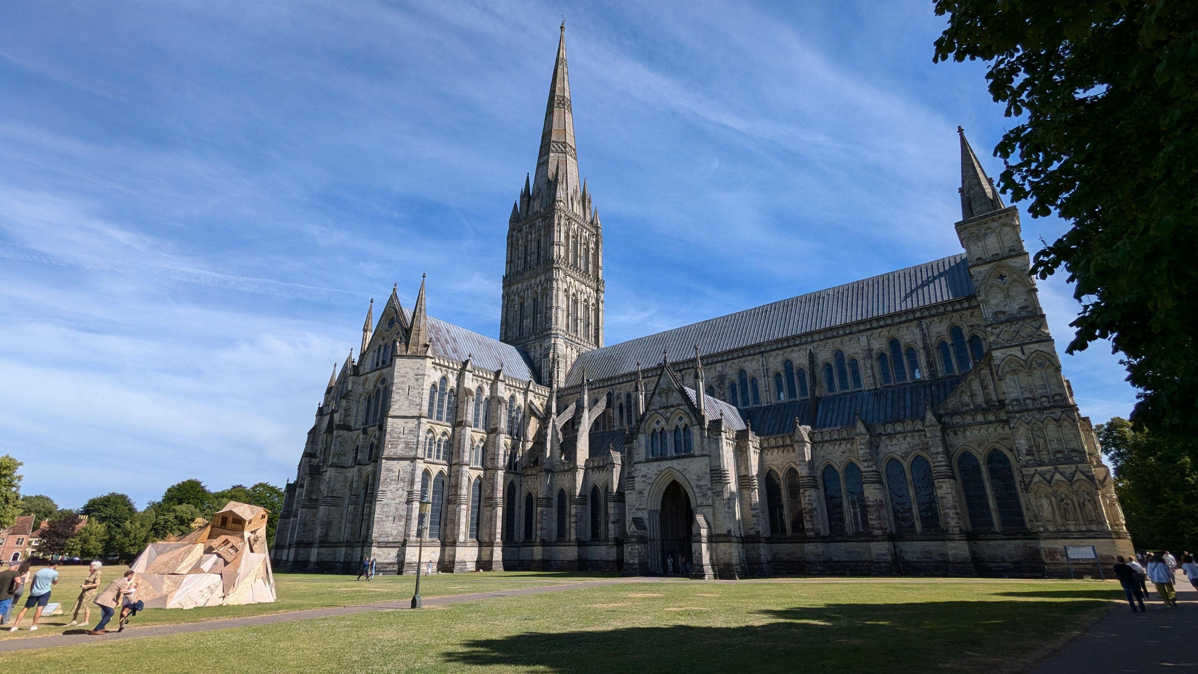 Looking up at Salisbury Cathedral from the side on a sunny day. Large, grand building with spire surrounded by green lawn