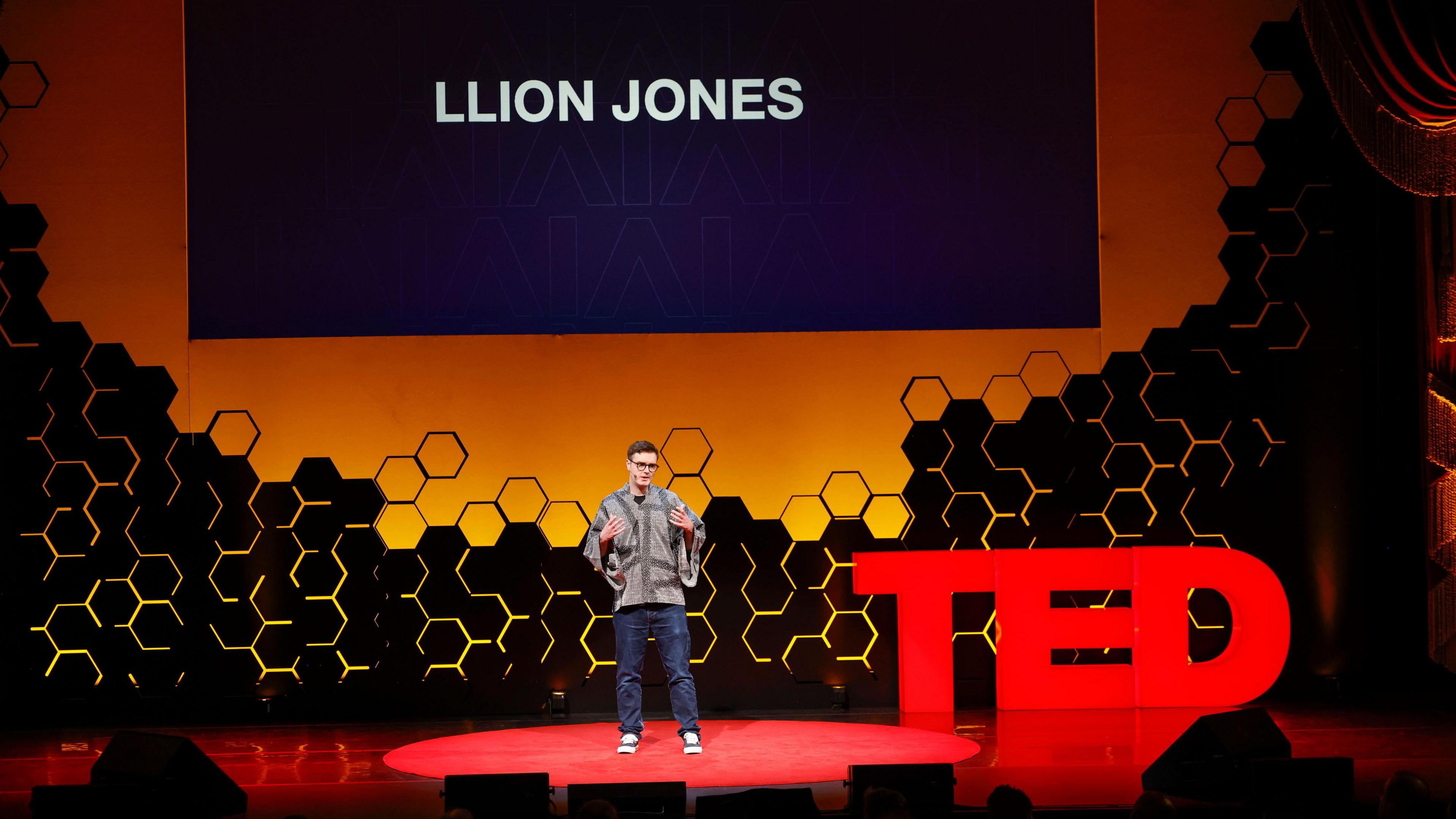 Llion Jones yn sefyll ar lwyfan gyda chefndir oren a phatrymau hecsagon du ar y wal. Mae’r person yn gwisgo crys â phatrwm llwyd tywyll a golau, a jîns glas tywyll gyda trainers gwyn. Mae’r llwyfan yn goch, ac mae’r enw “LLION JONES” wedi’i arddangos mewn llythrennau mawr ar y sgrin uwchben y siaradwr. 
