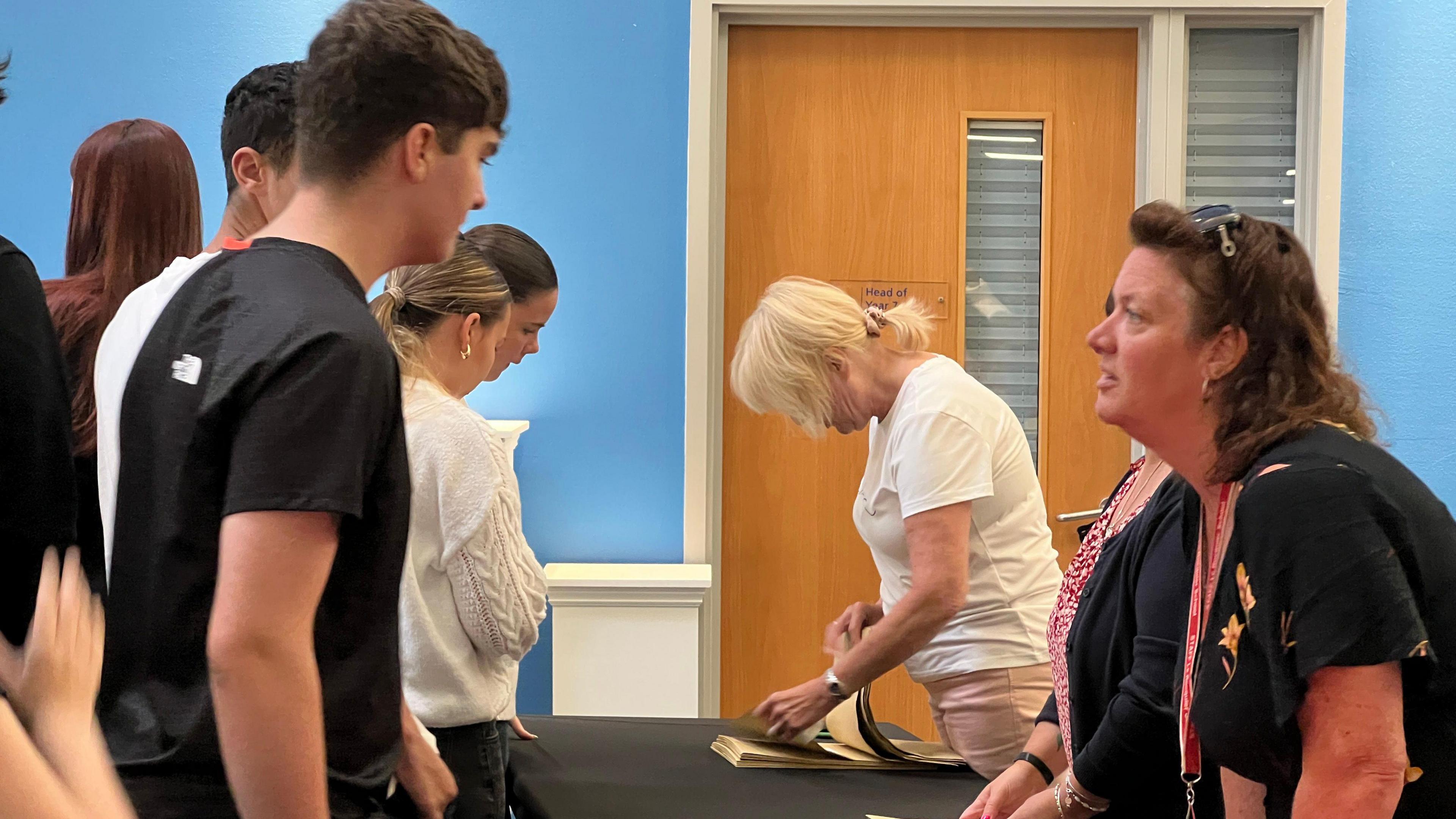 A group of people gathered around a table in a room with blue walls. Two individuals behind the table are distributing or organizing items, possibly books or documents. A wooden door with a small window and a sign is visible in the background.