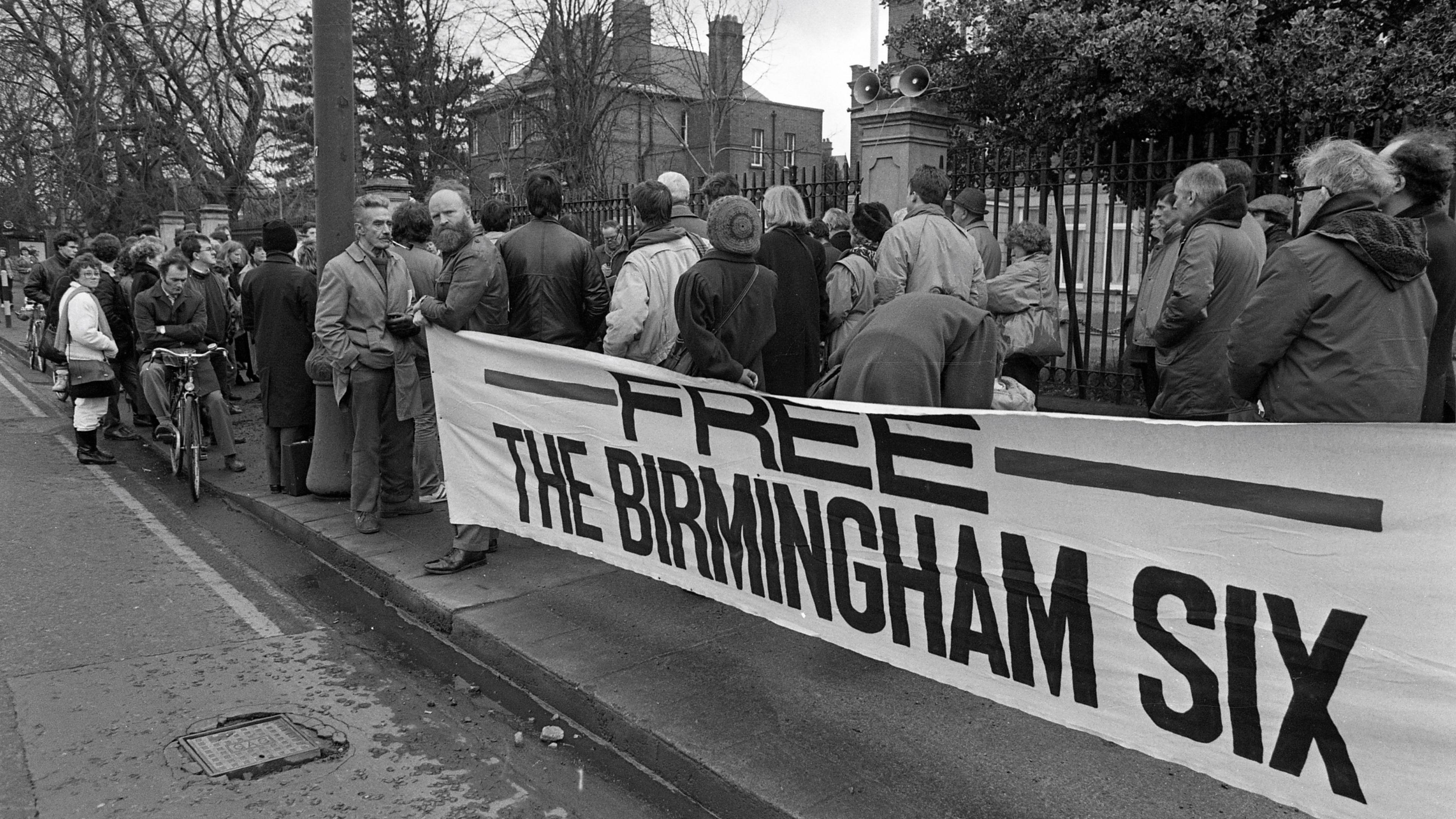 Black and white image of a group of people standing on a pavement in front of a large banner that says 'Free the Birmingham Six', with houses, a metal fence and trees behind them.