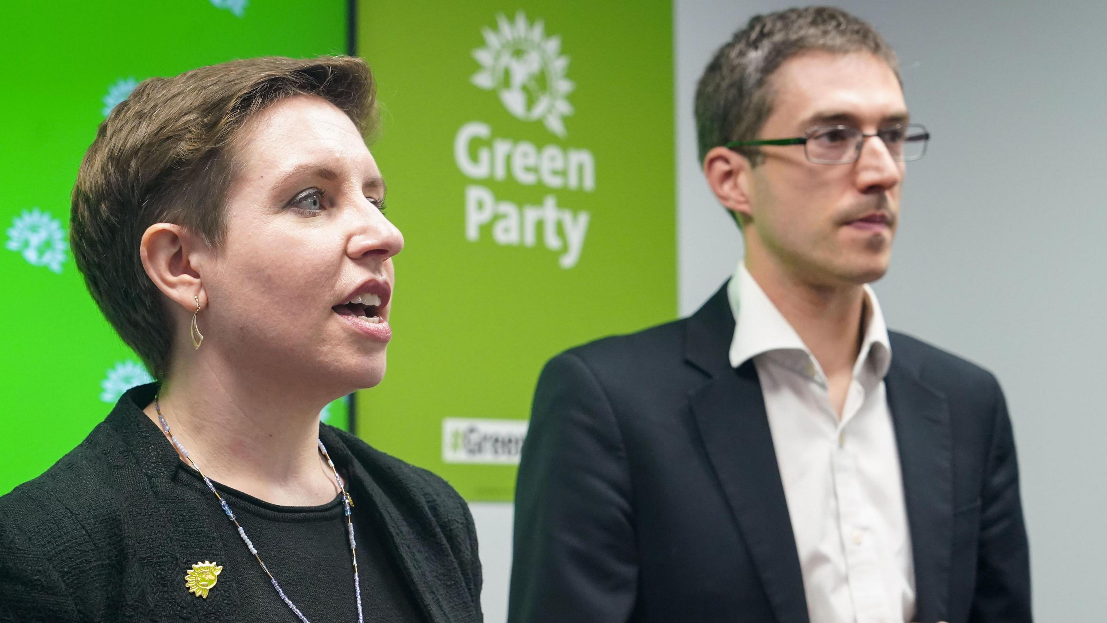 Carla Dener in a black jacket and t-shirt and Adrian Ramsau wearing a dark jecket and open necked white shirt stand on a stage with Green Party branding behind them