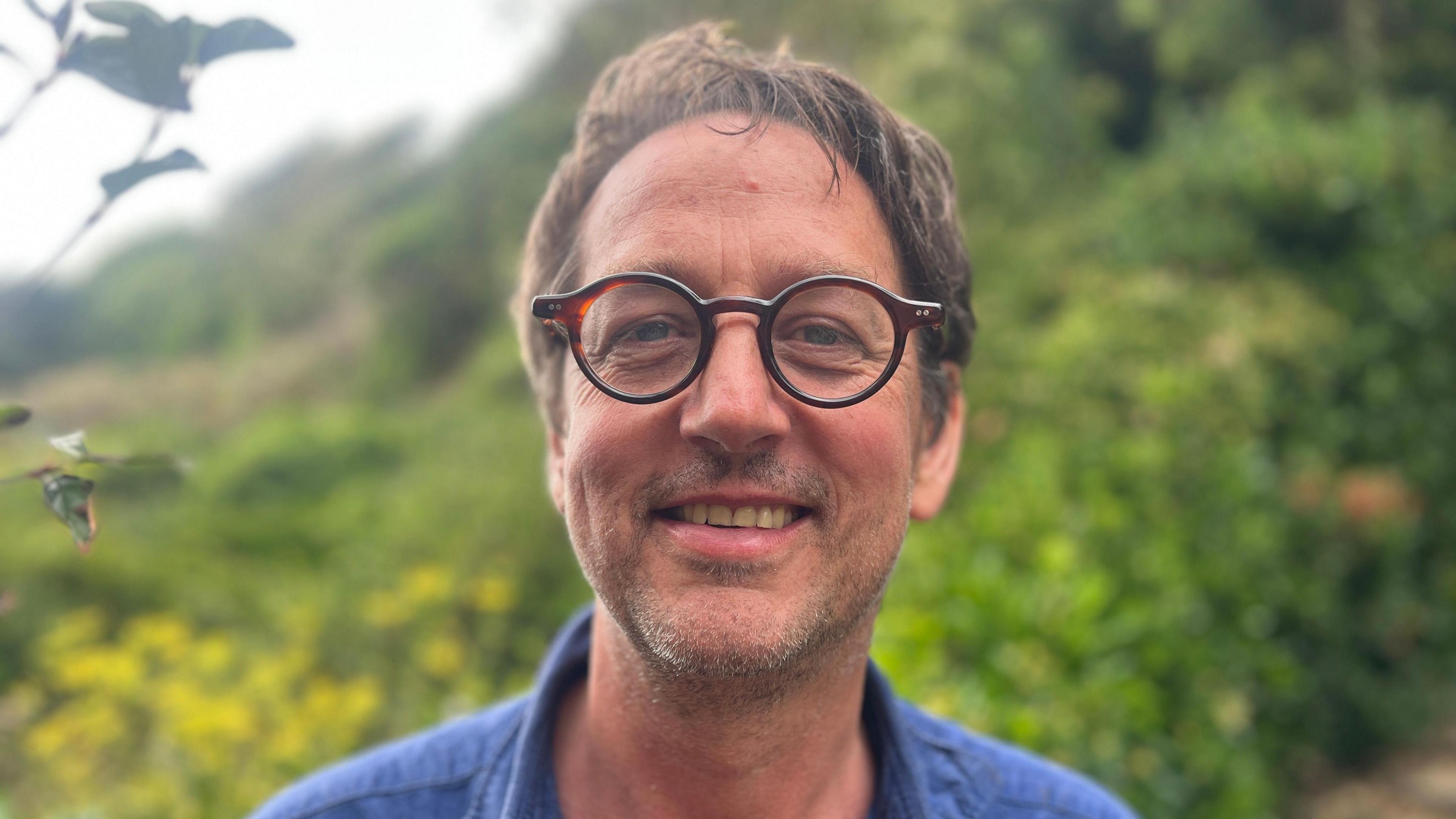Ben Ramsden smiles to the camera, standing in front of where the landslip happened. He wears a blue shirt and has short dark hair and glasses.