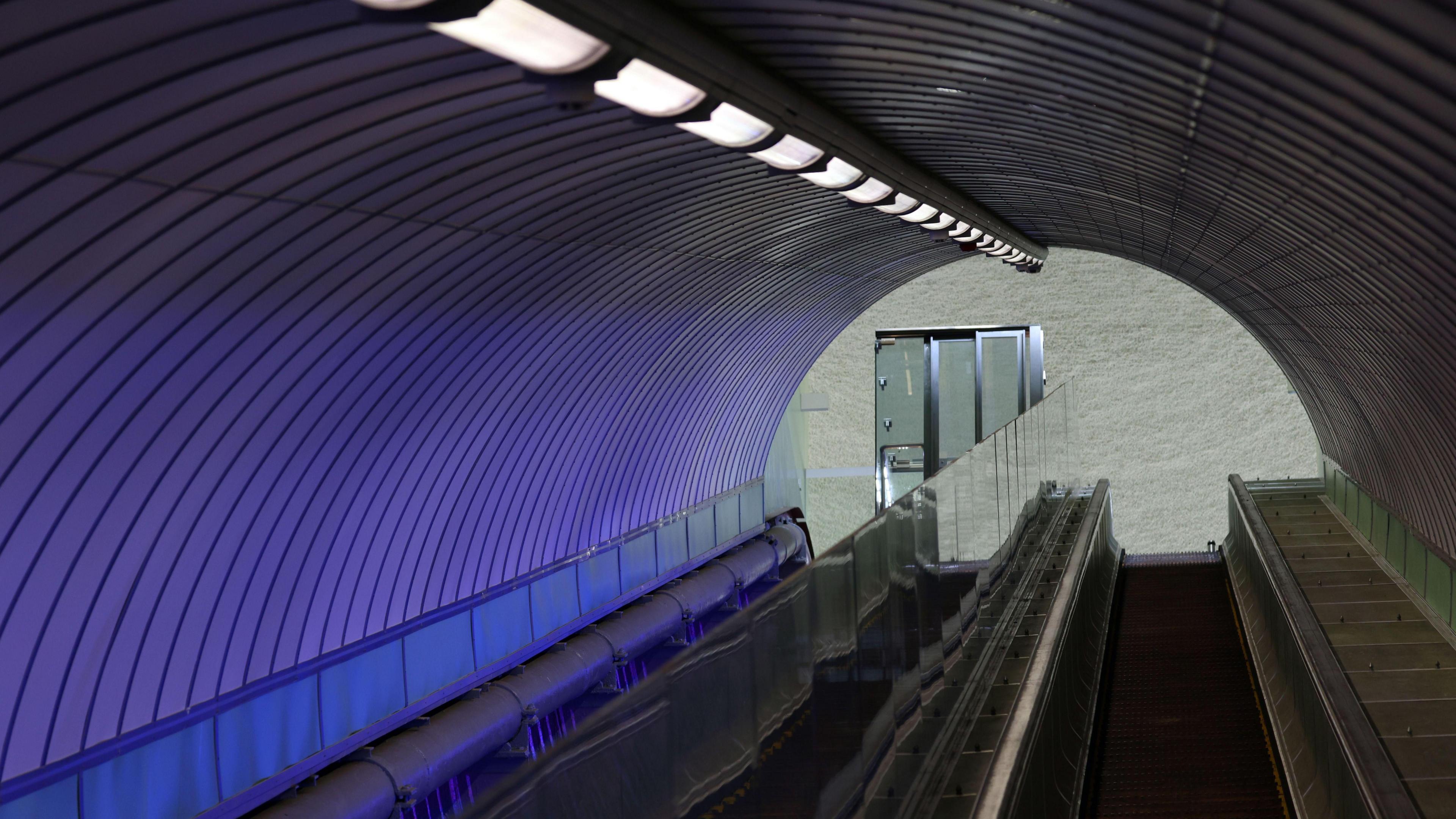 Looking up an inclined tunnel entrance covered in a blue light. Escalators can be seen running through it and there is a glass lift at the top.