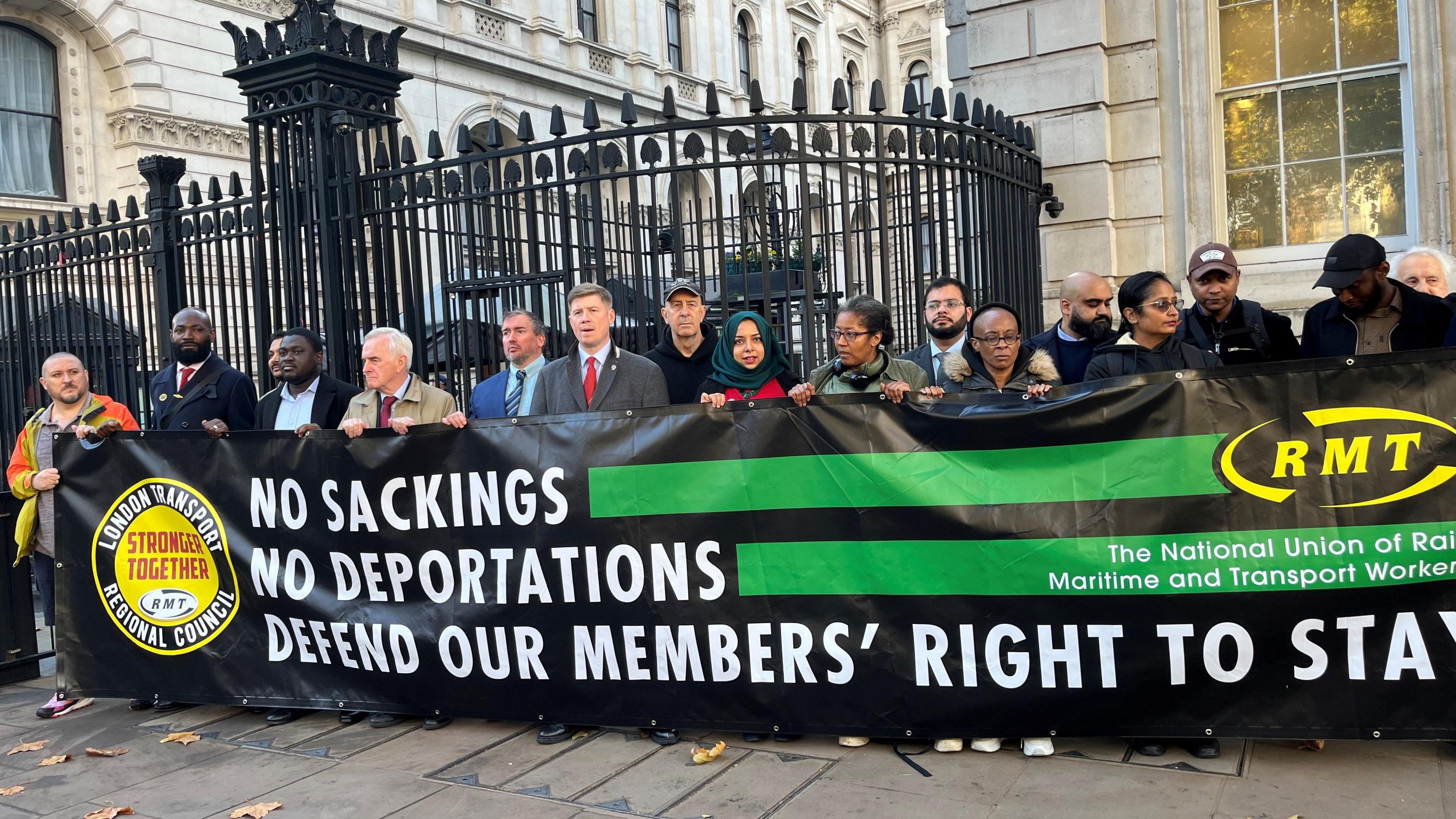 RMT general secretary Eddie Dempsey (centre) with workers holding a large black and green banner which reads "NO SACKINGS, NO DEPORTATIONS, DEFEND OUR MEMBERS' RIGHT TO STAY"