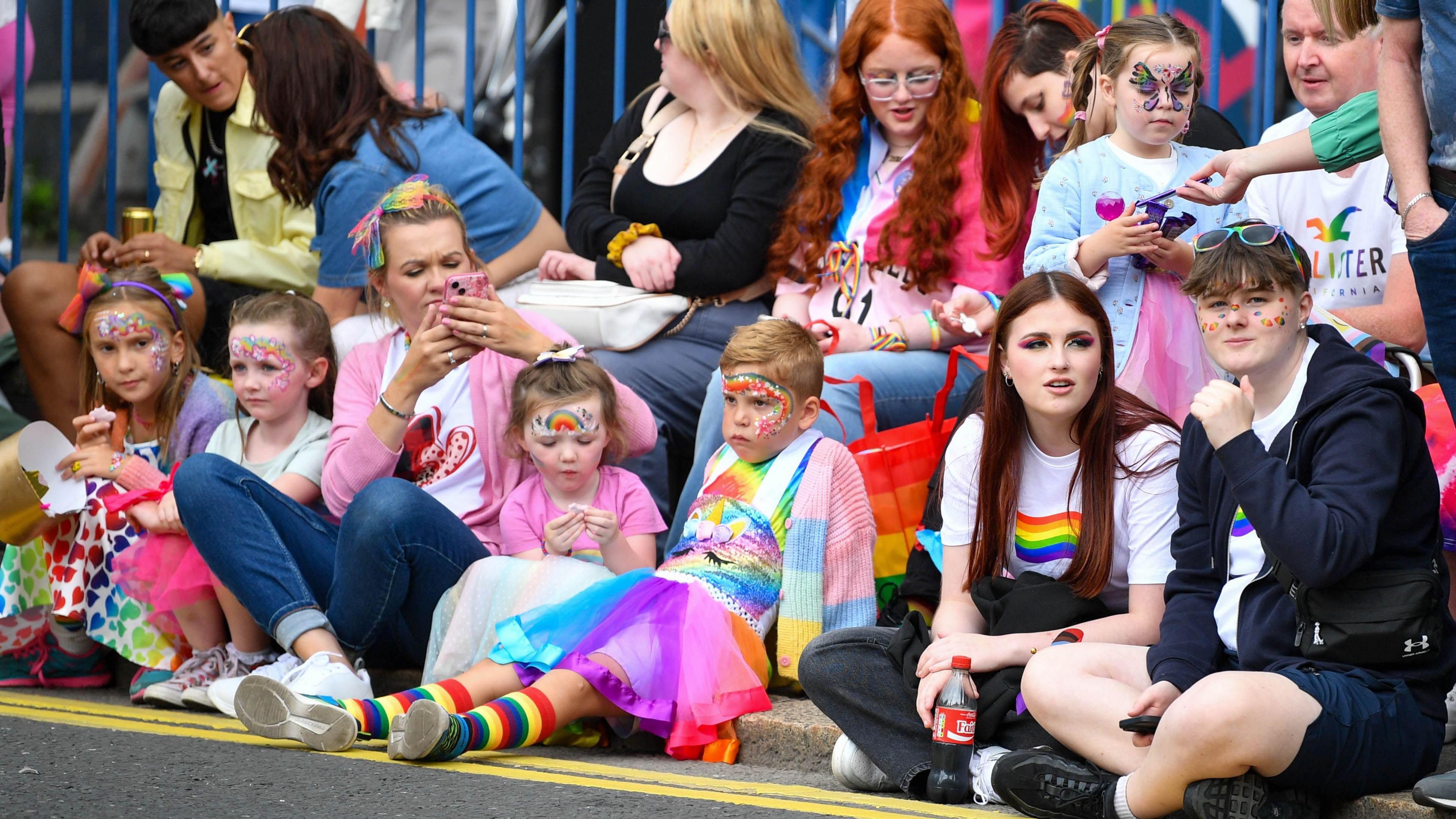 Spectators at a pride festival are sitting along a pavement. Children have theirs faces painted.