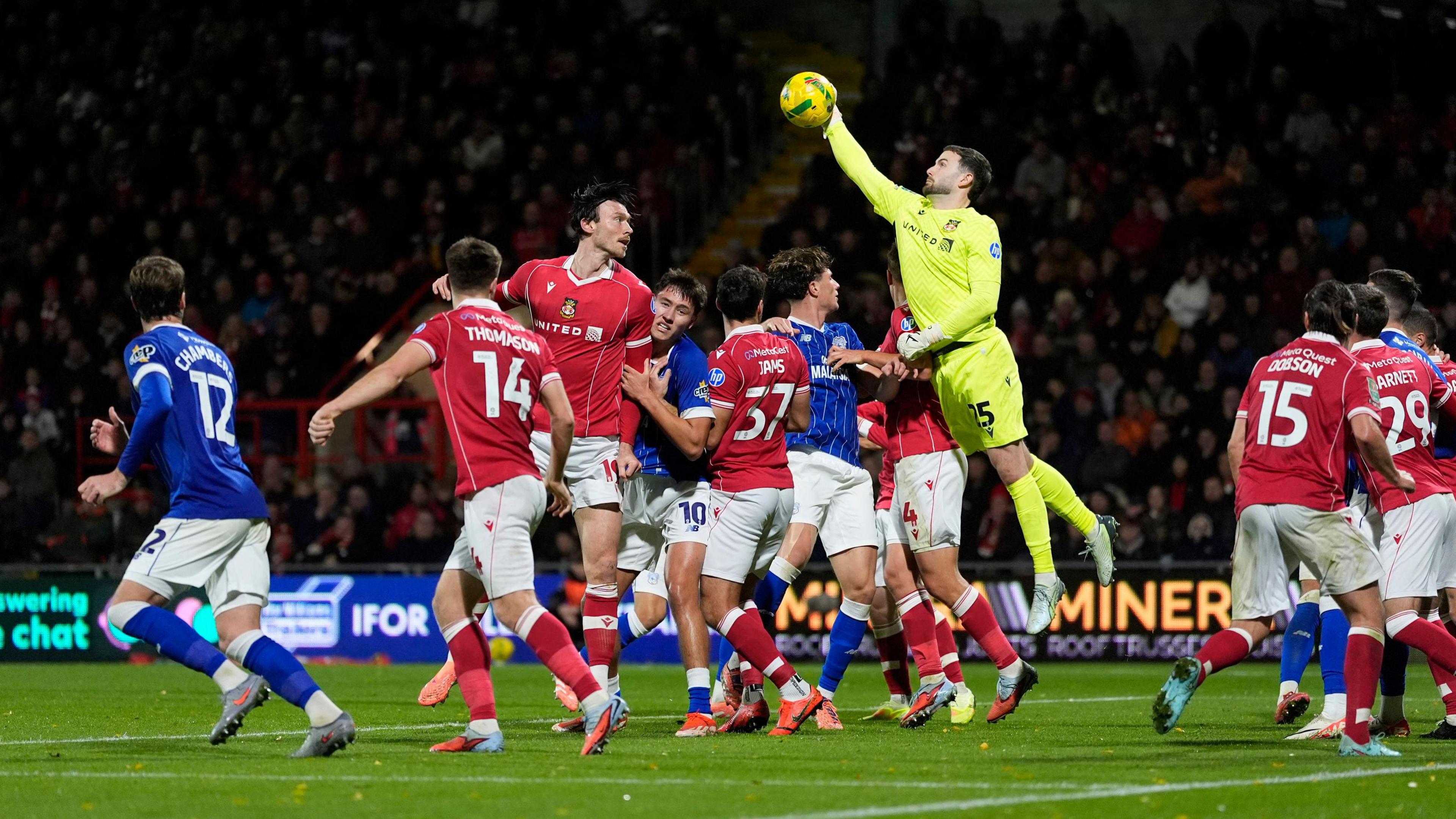 A goalkeeper is jumping high in the air to punch a football out of a box full of Wrexham AFC and Cardiff City players, sent in from a set-piece