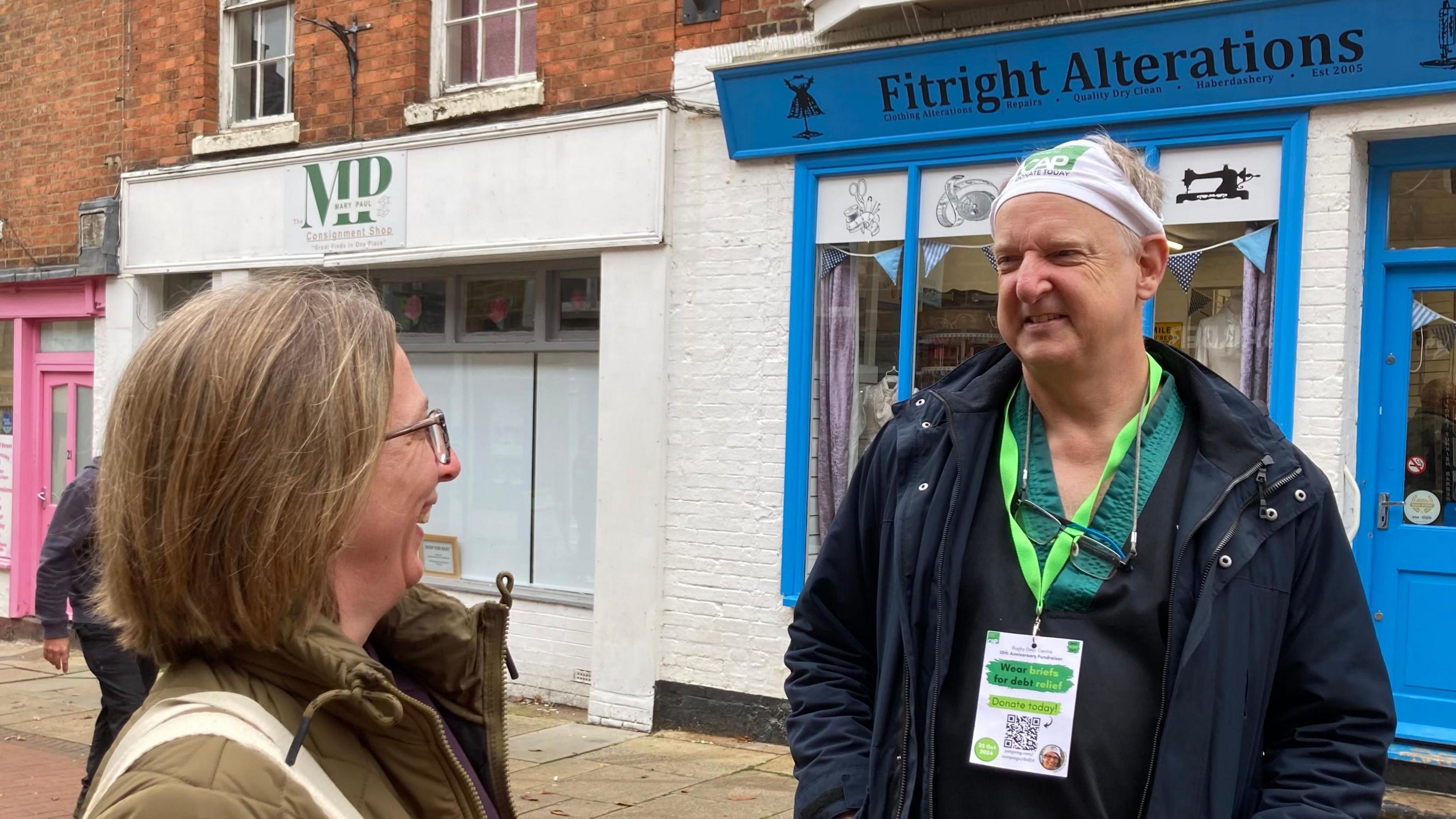 Woman with medium length brown hair and glasses wearing a green raincoat and carrying a cream-coloured handbag laughs at Mark Robson wearing his pants on his head in a shopping street. There is a shop with blue doors behind him.