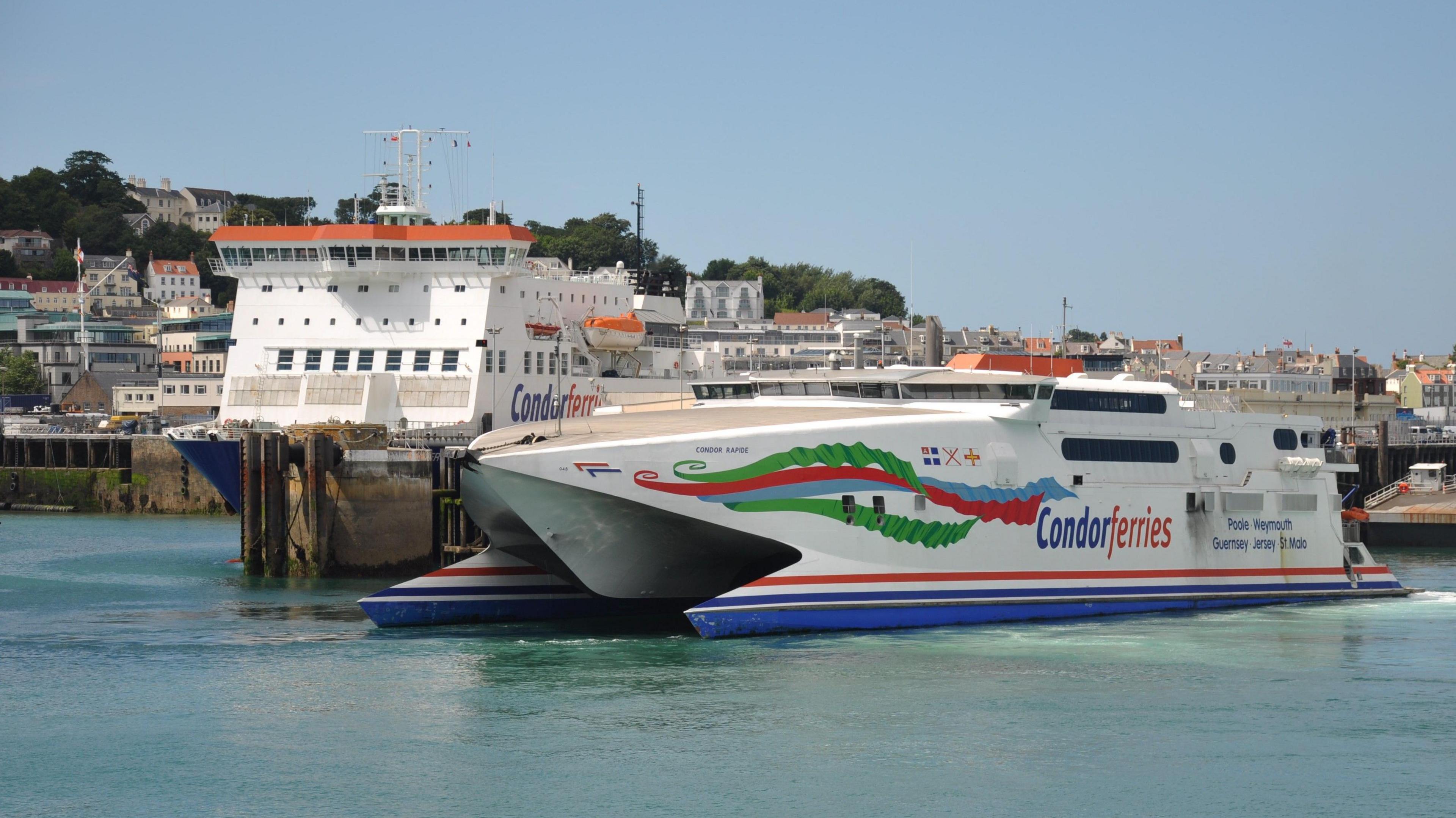 A white, blue, red and green ferry with Condor Ferries livery is docking in St Peter Port Harbour. Behind it you can see another ferry and some of the St Peter Port skyline.