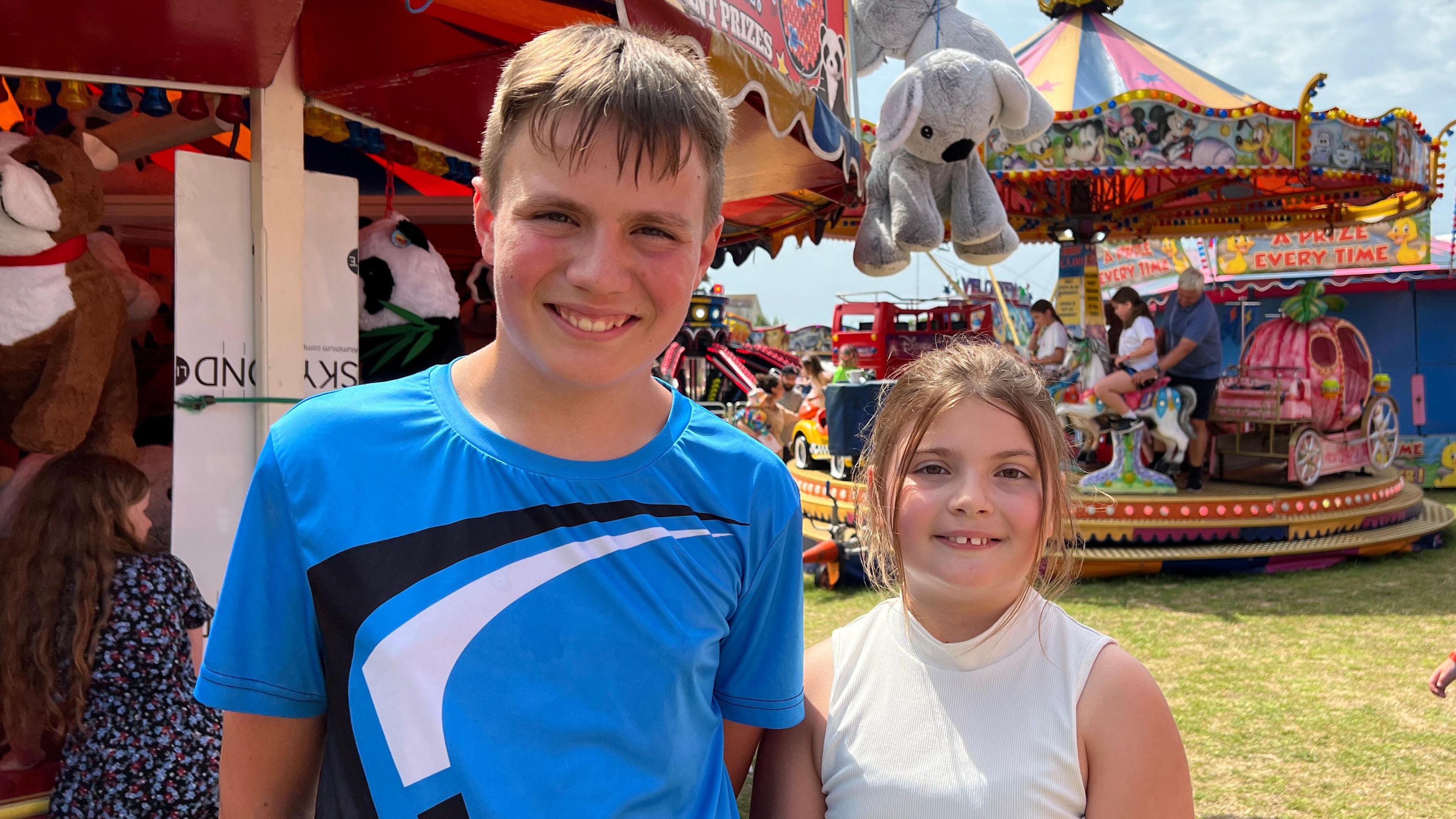 Max and Jessica smile at the camera with funfair rides behind them