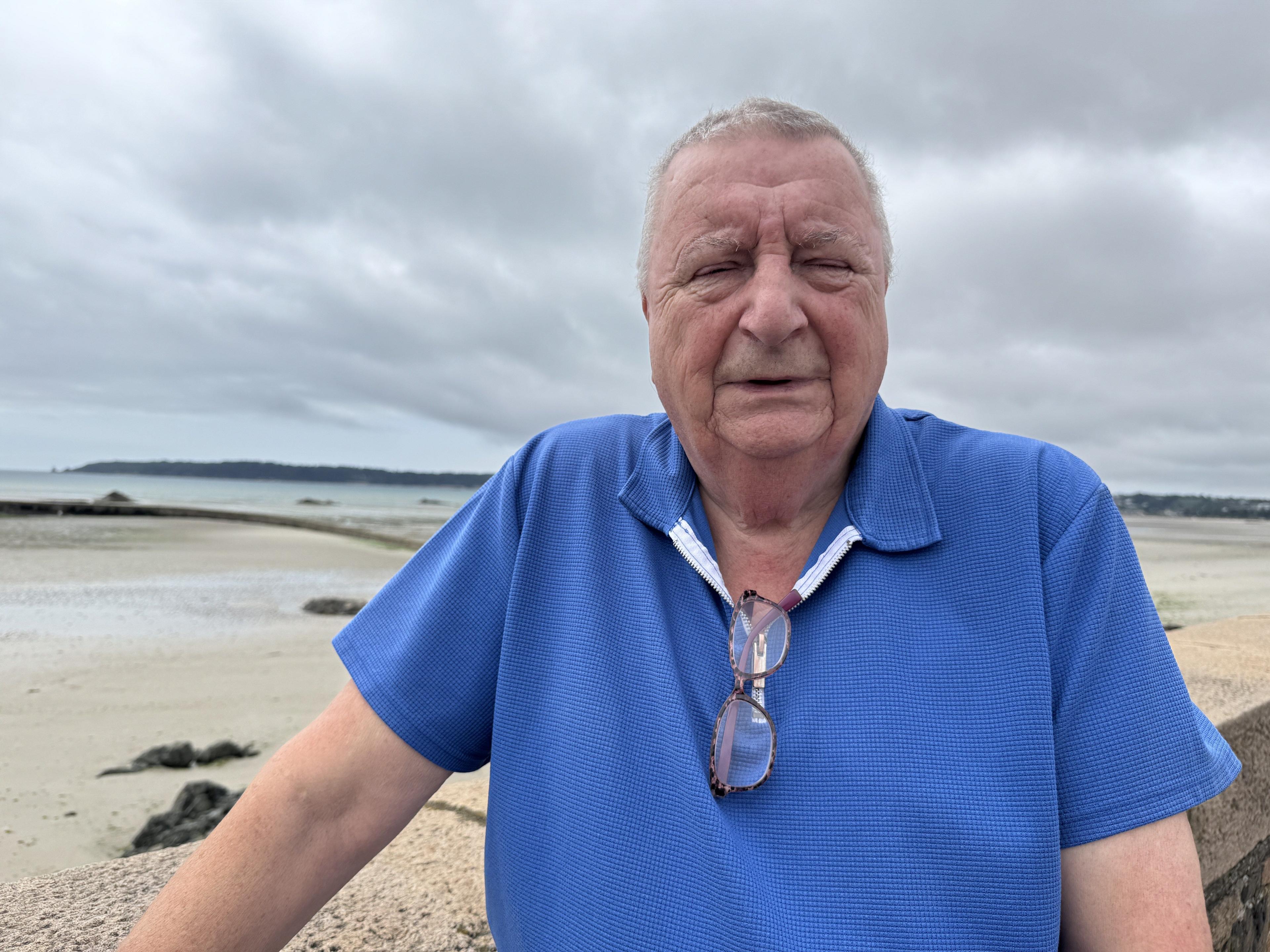 A man stands outdoors near a beach, wearing a blue short-sleeved shirt with glasses hanging from the collar. The background features a sandy beach, rocks, and an overcast sky.