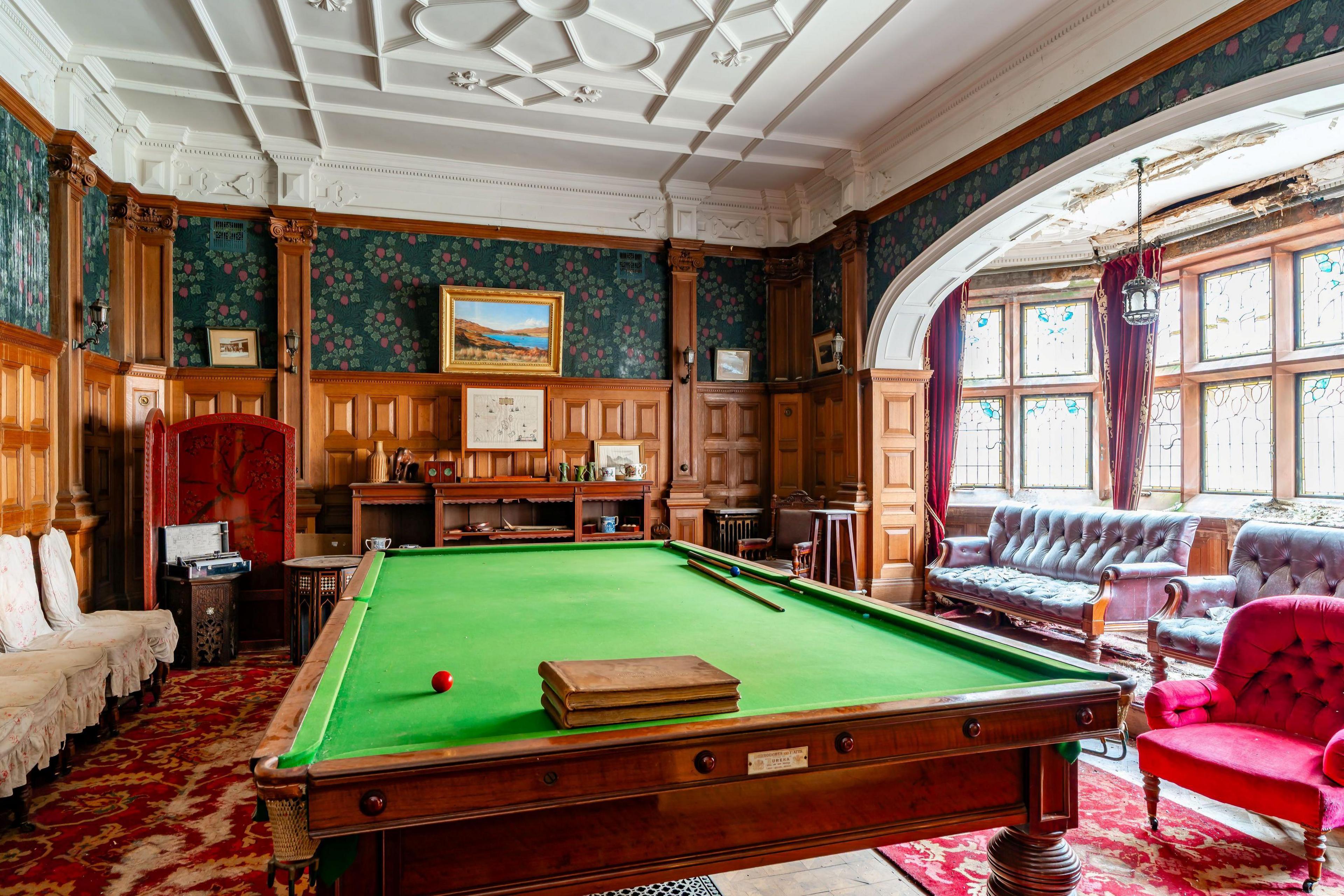An interior view of a room inside the KInloch Castle. There are large windows with ornate decorations in the glass. A billiard table fills most of the wood-panel walled room. 