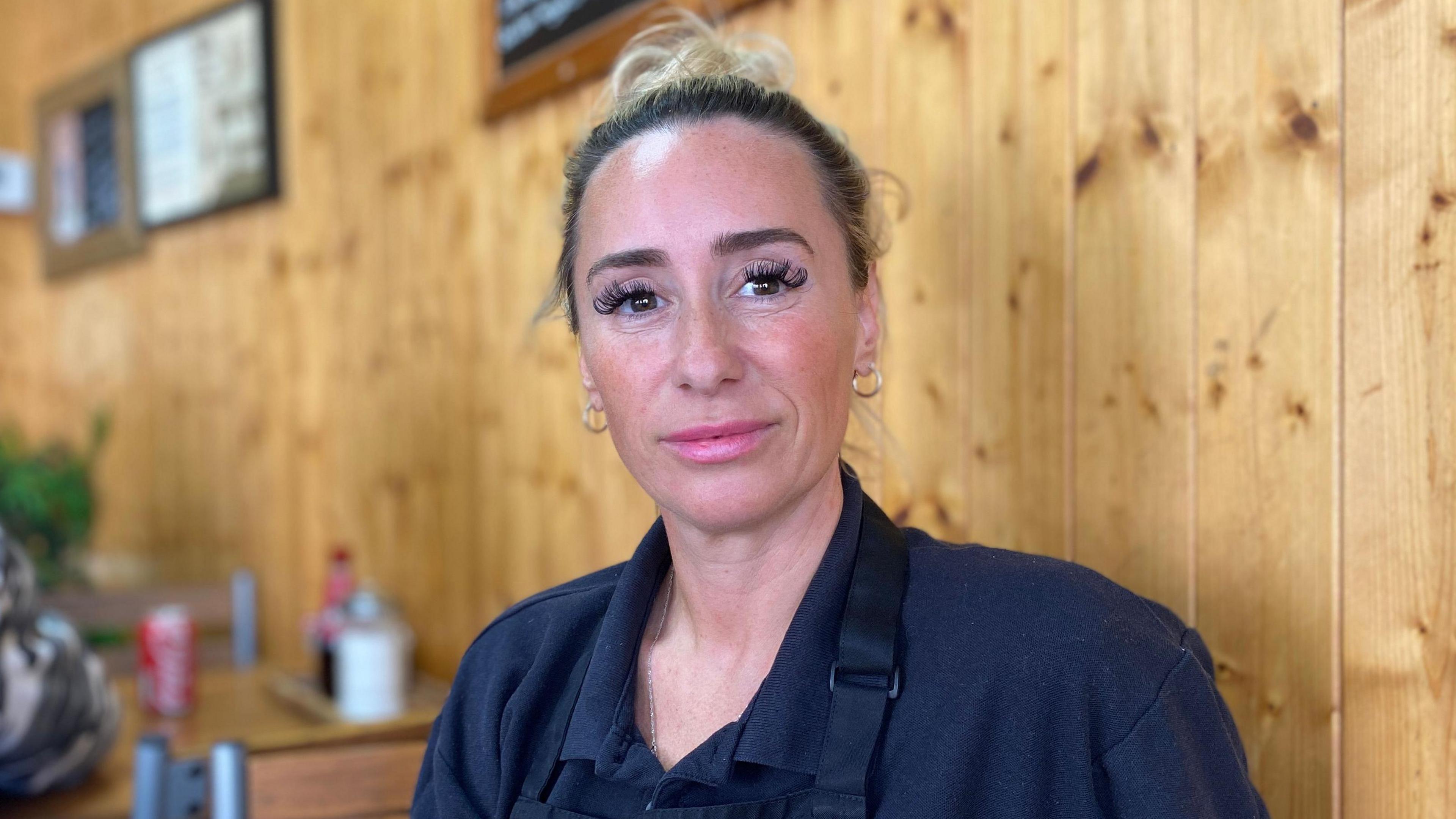 A woman has blonde hair tied in a bun and is wearing a navy blue shirt with a navy blue apron. She is sitting on a chair next to a wall with wooden panelling. There are tables with condiments on them behind.